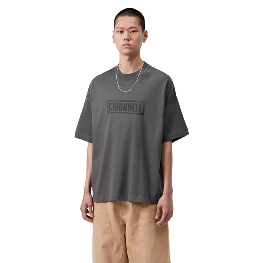 T-shirt Carhartt WIP S/S Bricky Man em Soot Stone Dyed — modelo masculino de frente, meio corpo, cabelo curto, brinco pequeno, fio de prata fino ao pescoço, gráfico Bricky Man impresso no peito: logotipo "Carhartt" em caixa retangular cinzento escuro tom-sobre-tom, malha de algodão orgânico cinzento Soot stone-dyed, calças cargo wide-leg bege Carhartt WIP, fundo branco.