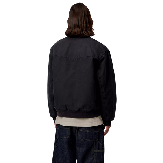 Casaco Carhartt WIP OG Santa Fe em Preto — modelo masculino de costas, meio corpo, lona Dearborn preta, costuras estruturais em arco no painel traseiro, faixa inferior em malha canelada preta, sweatshirt bege por baixo, calças cargo denim azul-escuro, fundo branco.