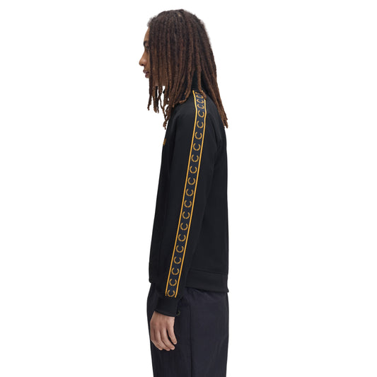Track jacket Fred Perry Contrast Tape Track Jacket em Navy — vista lateral com modelo, corpo inteiro, fita contrastante dourada com Laurel Wreaths repetidos ao longo da manga visível, combinado com calças navy Fred Perry.