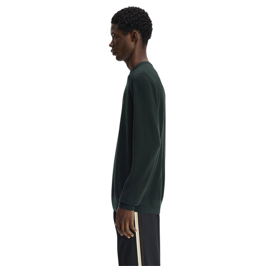 Camisola de malha para homem Fred Perry Pique Stitch Jumper em Night Green — modelo masculino de perfil direito, corpo inteiro, malha piqué verde-escuro Night Green, gola redonda canelada, punho canelado visível, regular fit, calças escuras com riscas laterais creme, fundo branco.