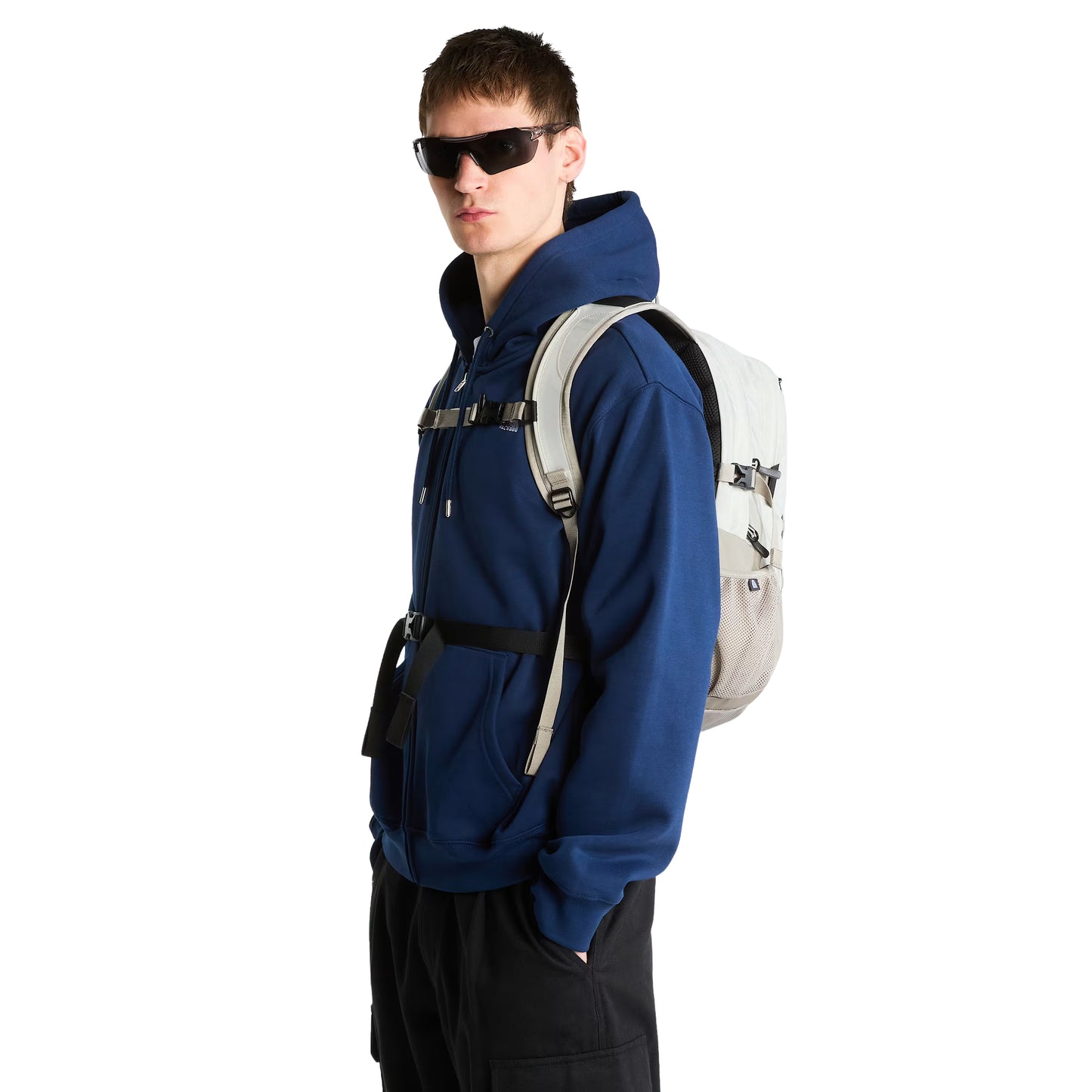 Mochila The North Face Borealis Classic em Cumulus Cloud / Stone Slab — modelo masculino de perfil a 3/4, hoodie azul marinho com capuz, calças pretas e óculos de sol escuros, mochila às costas com fivela de peito ajustada, alças FlexVent Stone Slab e bolso lateral em malha visíveis.