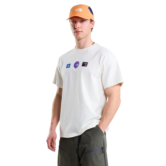 T-shirt unissexo The North Face Explorer Badges Relaxed em White Dune — modelo masculino de frente a 3/4, boné técnico laranja e azul TNF, calções cargo verde-oliva, três emblemas Explorer estampados em serigrafia à base de água no peito (azul retangular, roxo circular TNF, multicolor "1966"), corte relaxado com ombros descaídos visíveis, fundo branco.