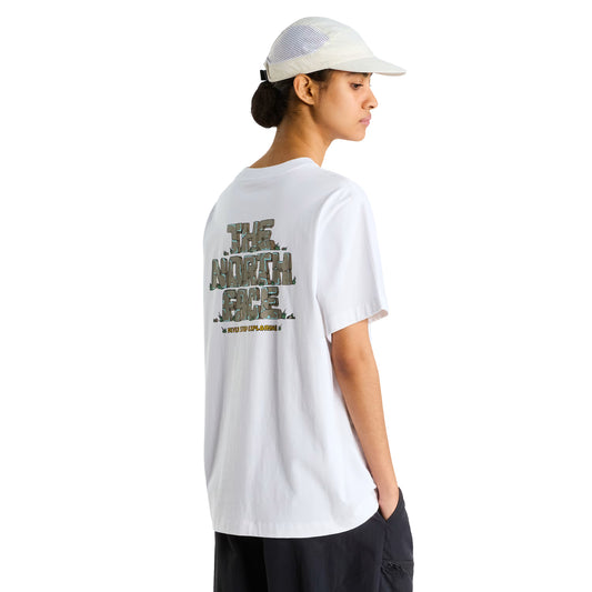 T-Shirt The North Face Stone Logo Relaxed em TNF White — modelo feminino de costas a 3/4, boné TNF branco perfurado, calças pretas cargo, gráfico "The North Face" estilo pedra em verde musgo/caqui e inscrição "Never Stop Exploring" em amarelo nas costas visíveis, fundo branco.