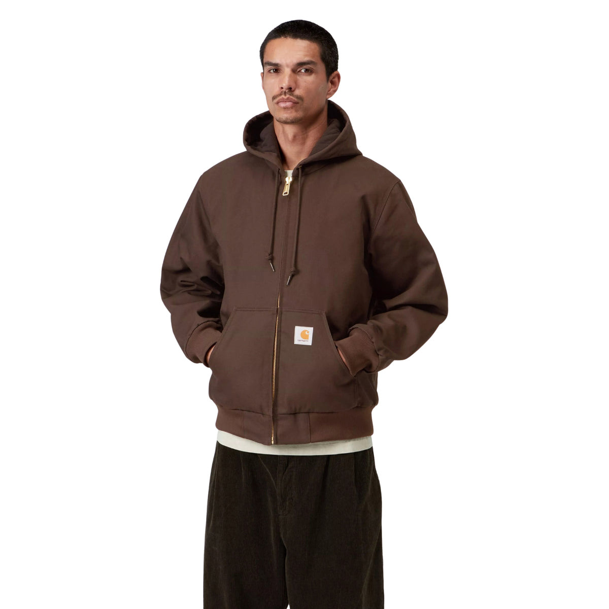 Casaco com carapuço Carhartt WIP Active em Tobacco Rigid, vista frontal em plano médio no modelo, com fecho fechado, mãos nos bolsos frontais, cordões do carapuço e etiqueta quadrada visível.
