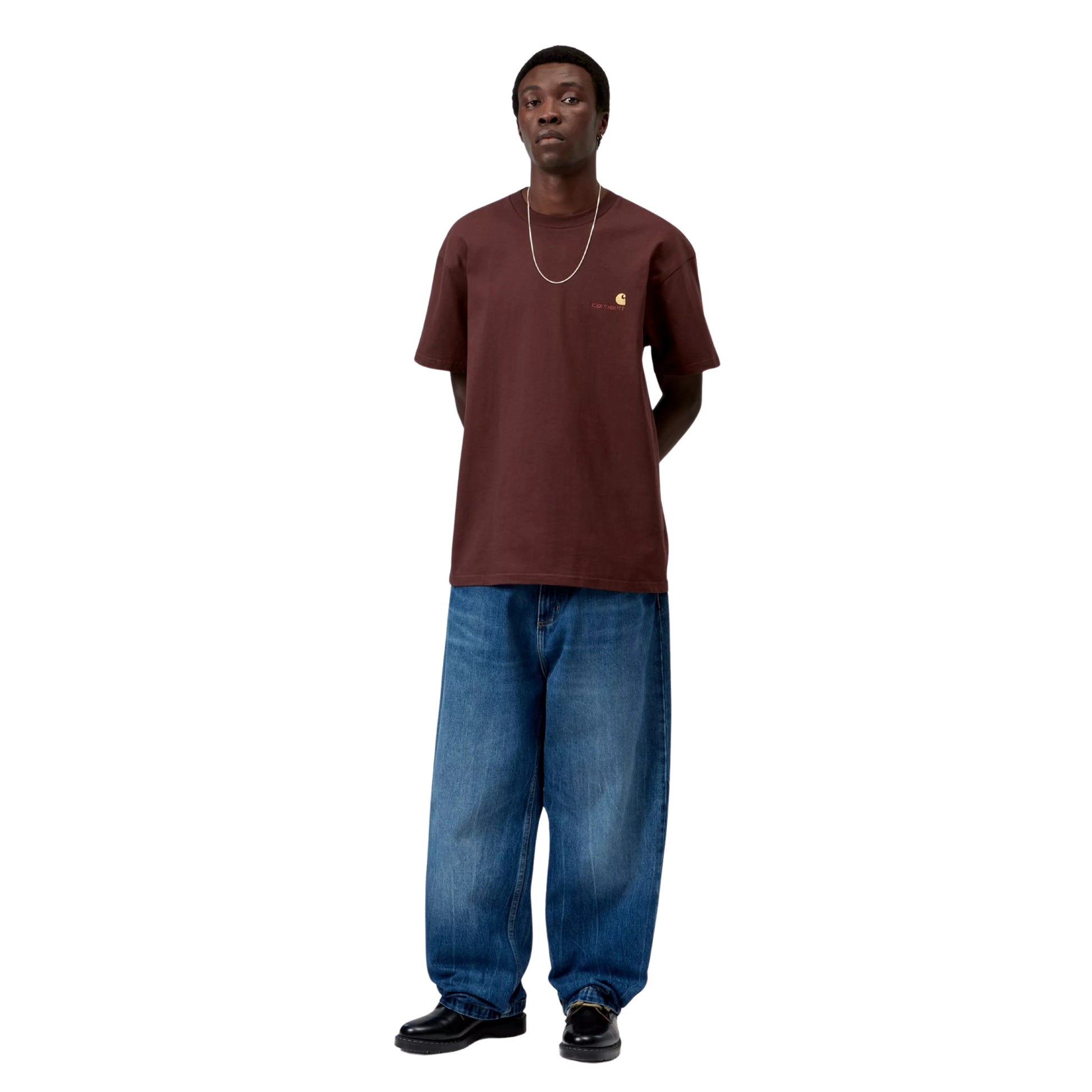 T-shirt Carhartt WIP American Script em Amarone — modelo masculino de frente, corpo inteiro, corrente fina dourada ao pescoço, logótipo American Script bordado no peito esquerdo com "C" dourado, calças wide-leg denim azul médio largas, loafers pretos, jersey de algodão bordeaux escuro Amarone, fundo branco.