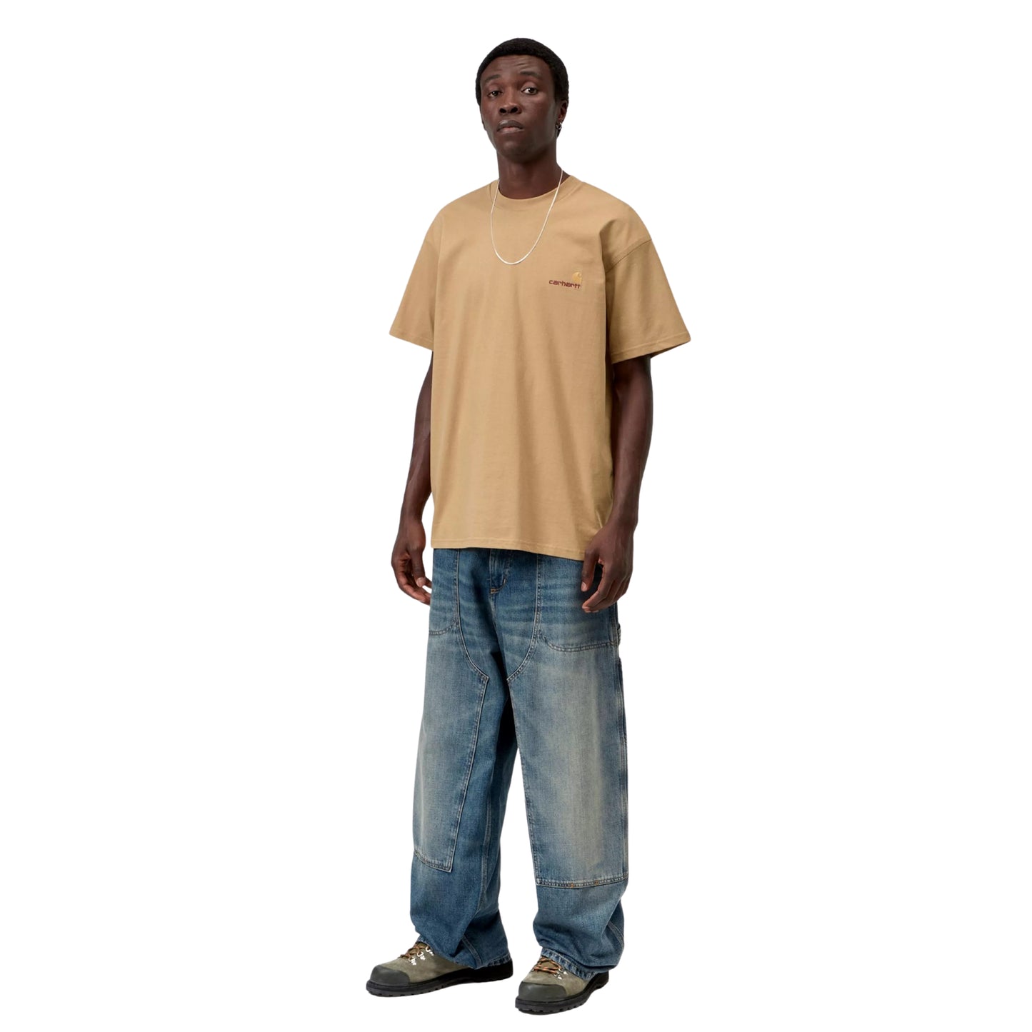 T-shirt Carhartt WIP S/S American Script em Dusty H Brown — modelo a usar a peça de frente, corpo inteiro, t-shirt bege-camel arenoso de corte solto, logótipo American Script de dois tons no peito esquerdo, calças Double Knee em ganga azul-médio desgastada e botas de caminhada cinzento-verde, fundo branco.