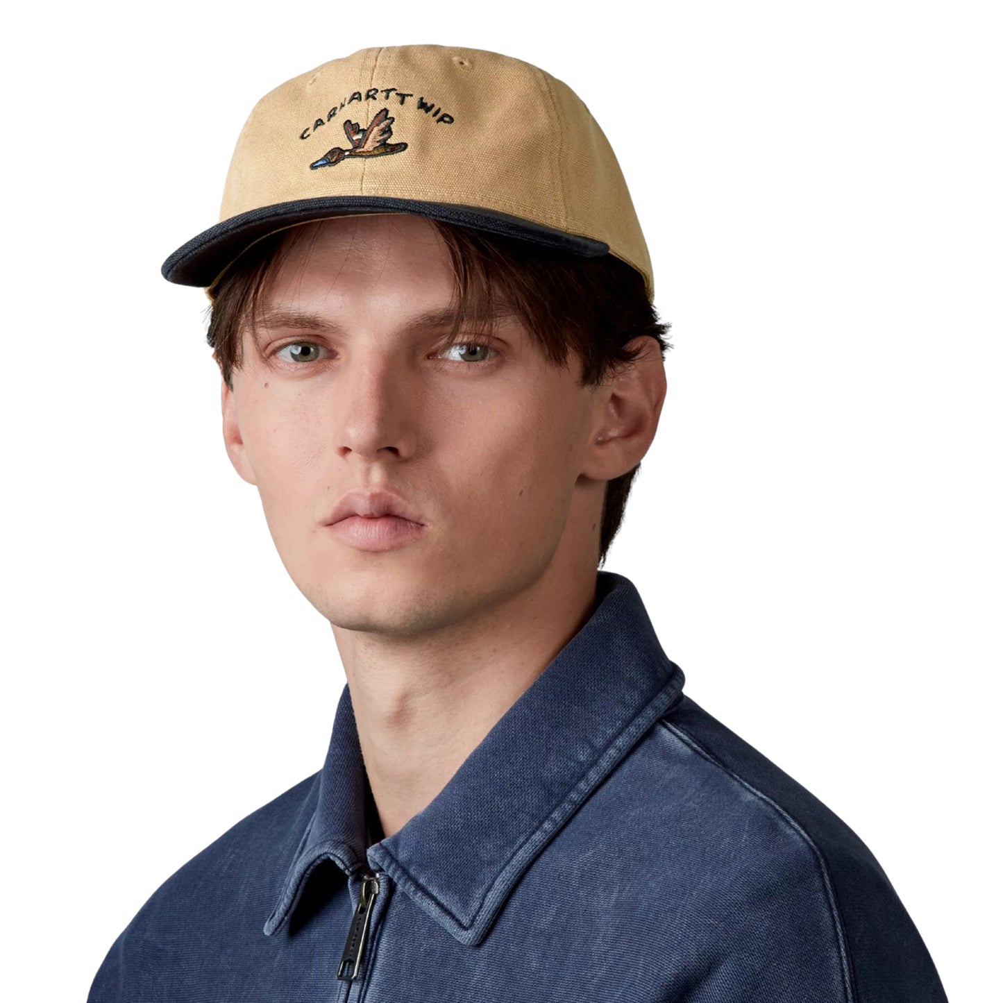 Boné Carhartt WIP Animated Ducks Cap em Dusty Hamilton Brown / Black — modelo masculino de frente, rosto, cabelo castanho curto, boné colocado na cabeça, bordado frontal com texto "CARHARTT WIP" em arco e pato animado pixel-art em voo em castanho e azul, copa não estruturada em lona Dearborn bege Dusty Hamilton Brown, pala ligeiramente curva preta, casaco de ganga azul-escuro por baixo, fundo branco.