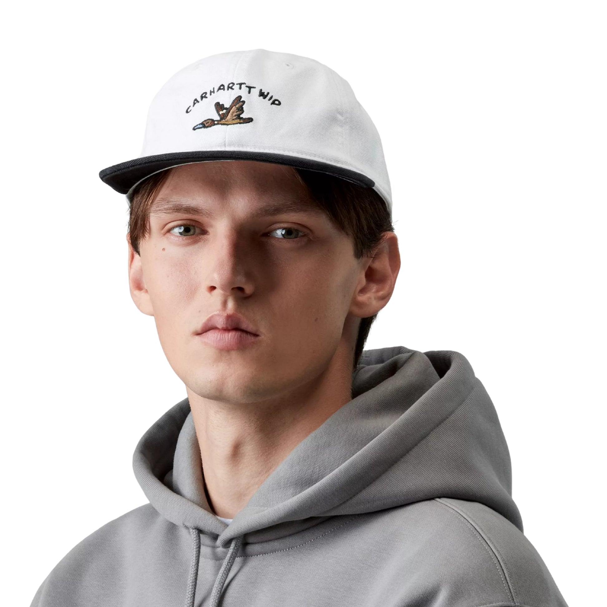 Boné Carhartt WIP Animated Ducks Cap em White / Black — modelo masculino de frente, rosto, cabelo castanho curto, boné colocado na cabeça, bordado frontal com texto "CARHARTT WIP" em arco em preto e pato animado pixel-art em voo em castanho e dourado, copa não estruturada em lona Dearborn branca, pala ligeiramente curva preta, sweatshirt com capuz cinzento por baixo, fundo branco.