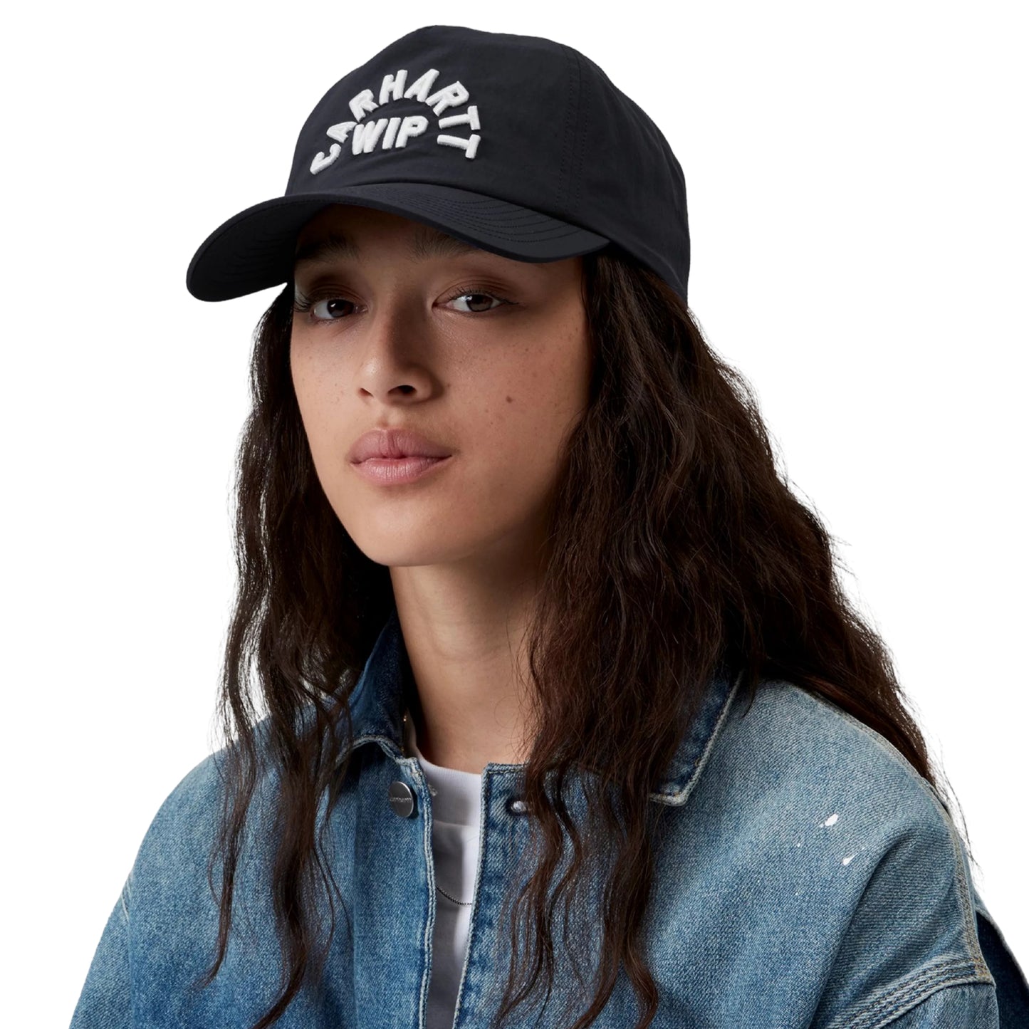 Boné Carhartt WIP Arch Script Cap em Dark Navy — modelo feminino de frente, meio corpo superior, cabelo castanho comprido ondulado solto, boné azul-marinho escuro de nylon sem estrutura com bordado tipográfico volumoso "CARHARTT WIP" em arco branco na frente, casaco de ganga azul-claro aberto, t-shirt branca interior, fundo branco.