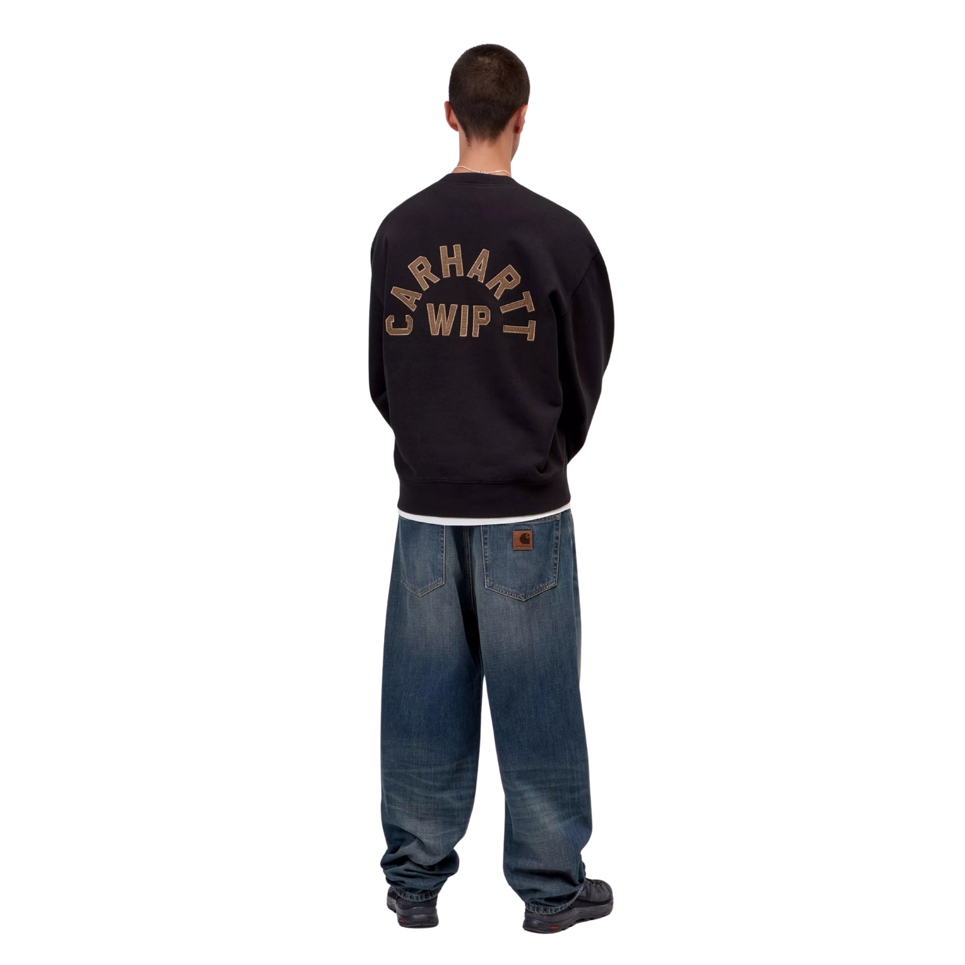 Sweat Carhartt WIP Arch Script em Black / Hamilton Brown Stone Washed — modelo masculino de costas, corpo inteiro, calças Carhartt WIP largas em denim azul lavado com patch castanho, sapatilhas pretas, bordado em arco "Carhartt WIP" em Hamilton Brown nas costas e corte balloon com bainha canelada visíveis, fundo branco.