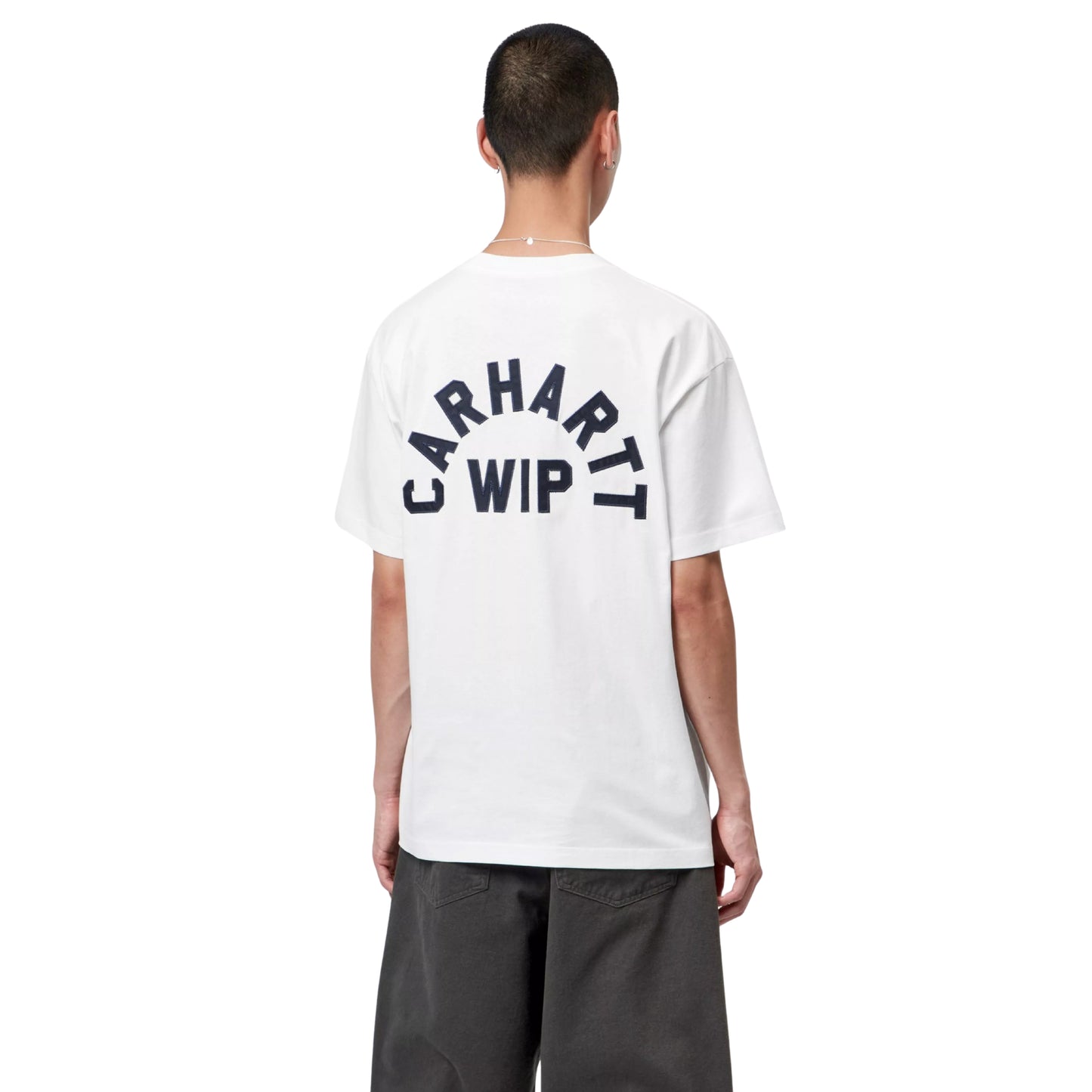 T-shirt Carhartt WIP S/S Arch Script em White / Dark Navy — modelo masculino de costas, meio corpo superior, cabelo curto, fio prateado ao pescoço, t-shirt branca de algodão orgânico com apliques tipográficos "CARHARTT WIP" em arco azul-marinho escuro nas costas, letras em relevo costuradas em jersey, calças wide-leg cinzento-escuras, fundo branco.