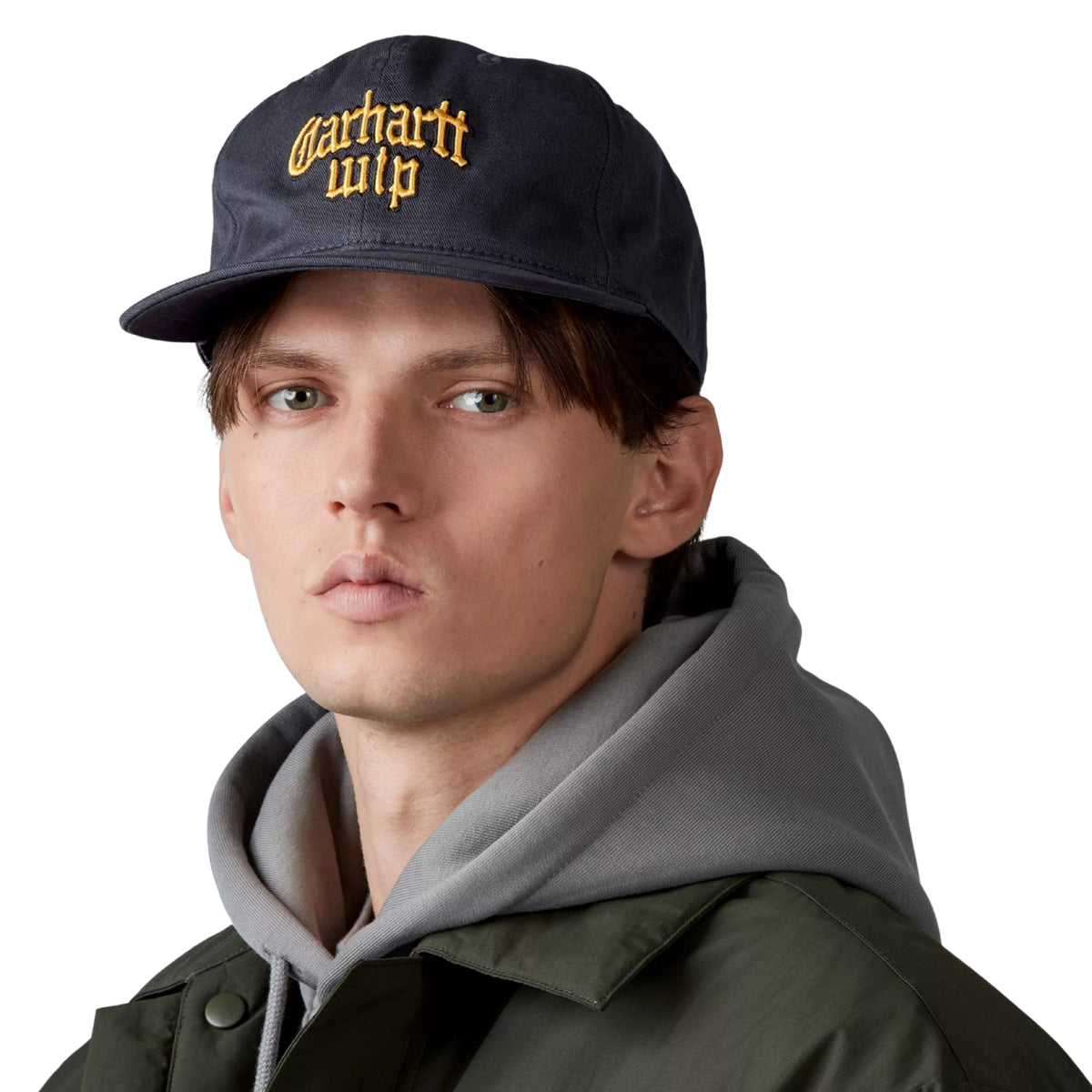 Boné Carhartt WIP Blackletter Cap em Dark Navy — modelo masculino de frente, meio corpo, boné six-panel azul-marinho com bordado Carhartt WIP em tipografia blackletter a dourado, pala plana, sweatshirt cinzenta e casaco verde por baixo, fundo branco.