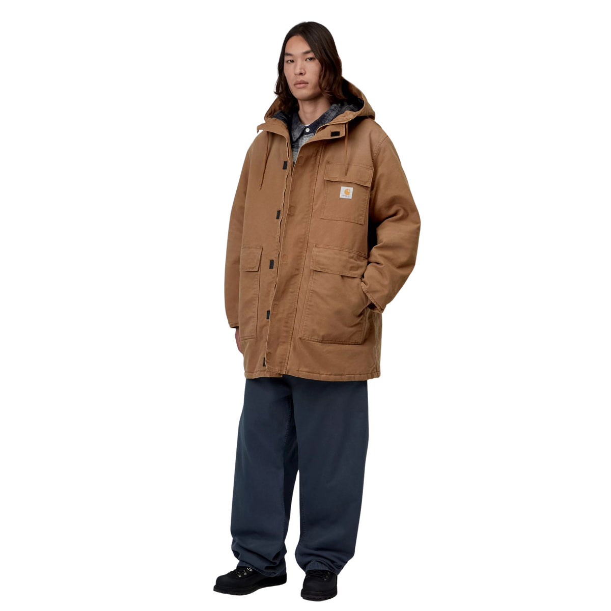 Parka Carhartt WIP Boreal em WIP H Brown — modelo masculino de frente, três quartos, fecho de correr aberto com sweatshirt xadrez cinzenta visível por baixo, bolso de peito e dois bolsos frontais com patilha, logótipo Carhartt WIP, calças largas azul-marinho, botas pretas, fundo branco.