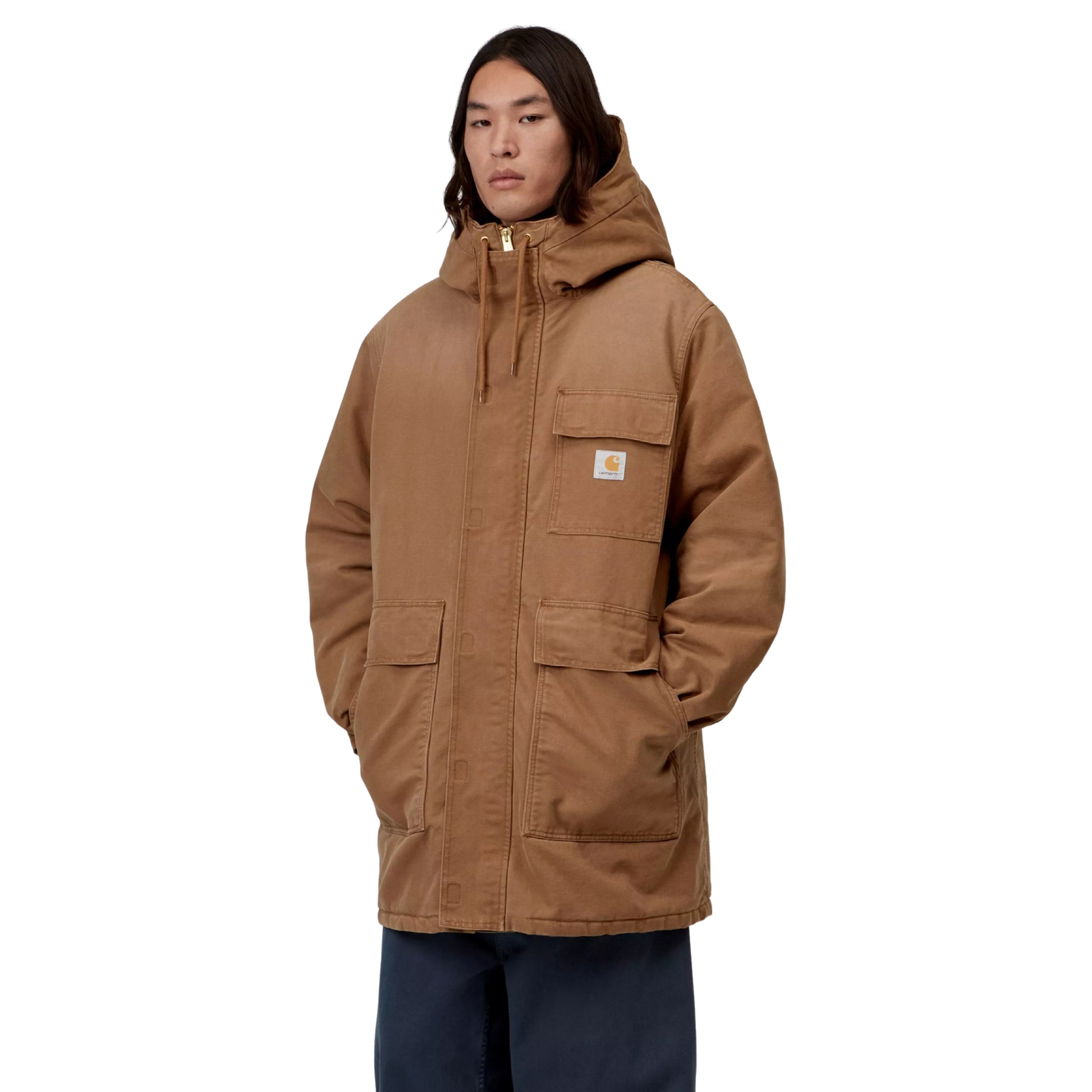 Parka Carhartt WIP Boreal em WIP H Brown — modelo masculino de frente, corpo inteiro, fecho de correr oculto fechado com puxador dourado, bolso de peito com patilha e logótipo Carhartt WIP, dois bolsos frontais de chapa, mãos nos bolsos, capuz com cordão, calças largas azul-marinho, fundo branco.