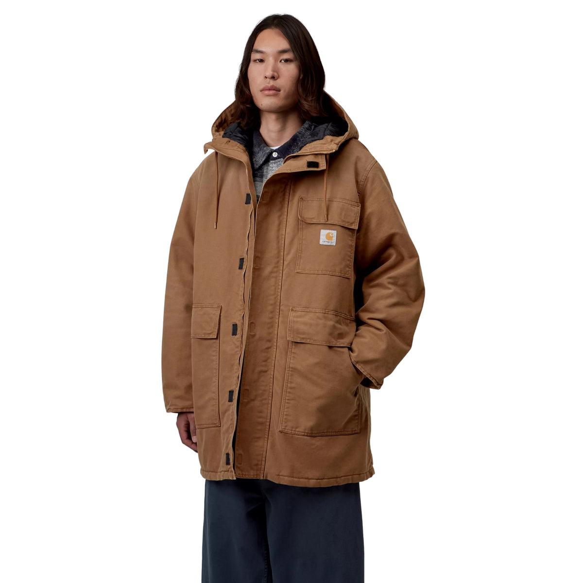 Parka Carhartt WIP Boreal em h brown stone canvas — modelo masculino de frente, corpo inteiro, fecho de correr aberto com sweatshirt xadrez azul visível por baixo, dois bolsos frontais de chapa e bolso de peito, logótipo Carhartt WIP, calças largas azul-marinho, fundo branco.