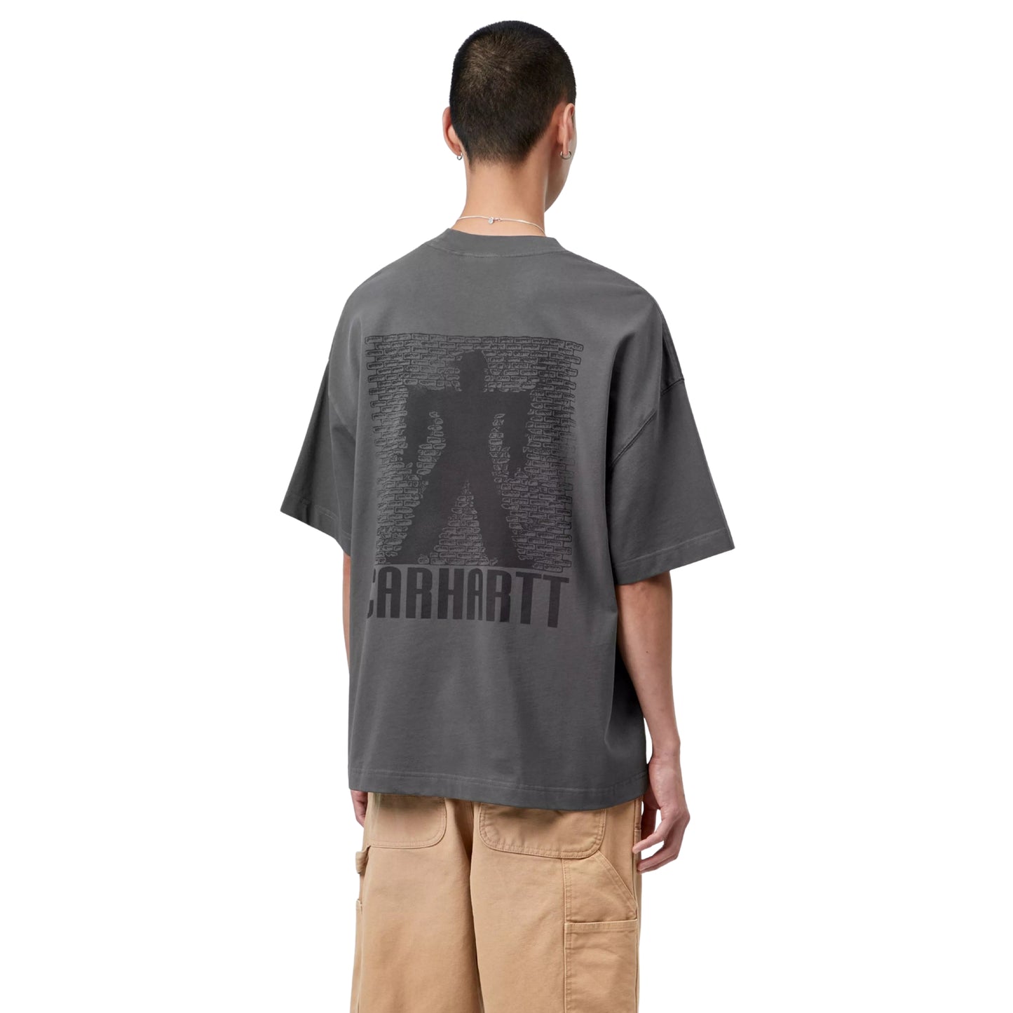T-shirt Carhartt WIP S/S Bricky Man em Soot Stone Dyed — modelo masculino de costas, meio corpo, cabelo curto, brinco pequeno, gráfico Bricky Man impresso nas costas: silhueta de figura humana de pé contra parede de tijolos em cinzento escuro tom-sobre-tom, texto "Carhartt" em baixo no mesmo tom, malha de algodão orgânico cinzento Soot stone-dyed, calças cargo wide-leg bege Carhartt WIP, fundo branco.