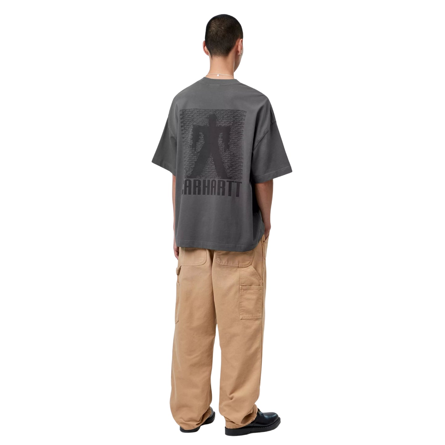 T-shirt Carhartt WIP S/S Bricky Man em Soot Stone Dyed — modelo masculino de costas, corpo inteiro, cabelo curto, brinco e fio visíveis, gráfico Bricky Man nas costas: silhueta humana contra parede de tijolos e texto "Carhartt" em cinzento escuro tom-sobre-tom, malha de algodão orgânico cinzento Soot stone-dyed, calças cargo wide-leg bege Carhartt WIP, sapatos pretos de sola grossa, fundo branco.