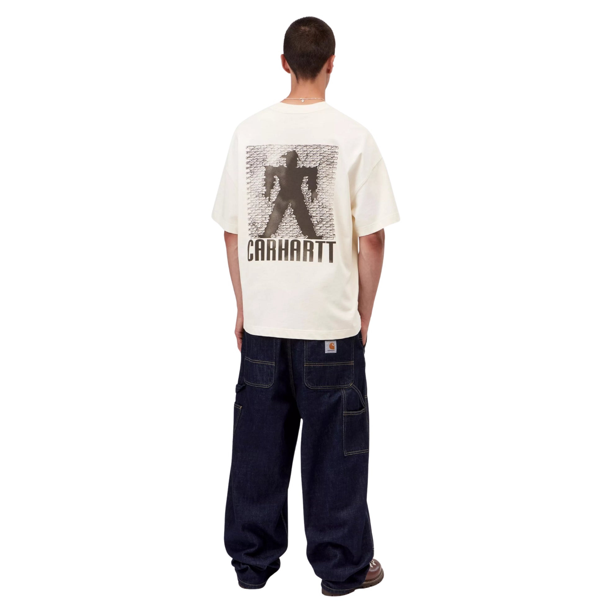 T-shirt Carhartt WIP S/S Bricky Man em Wax Stone Dyed — modelo masculino de costas, corpo inteiro, cabelo curto, gráfico Bricky Man nas costas com silhueta a preto e texto "Carhartt" em grande, malha de algodão orgânico bege creme Wax stone-dyed, calças wide-leg denim azul-escuro Carhartt WIP com costuras amarelas, sapatos pretos de sola grossa, fundo branco.