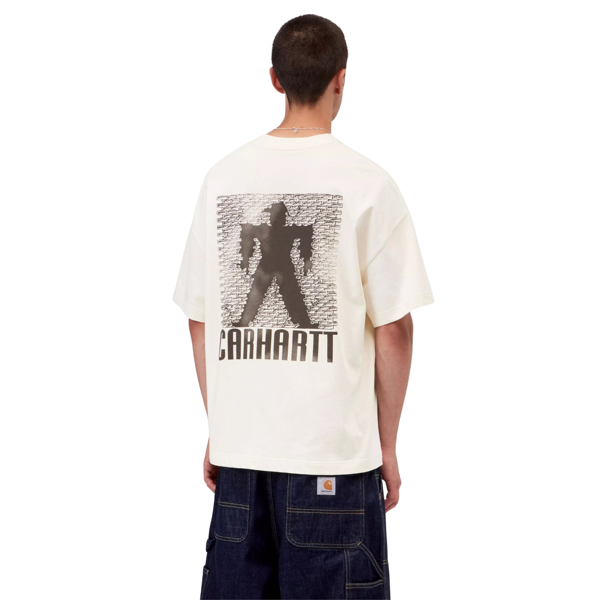 T-shirt Carhartt WIP S/S Bricky Man em Wax Stone Dyed — modelo masculino de costas, meio corpo, cabelo curto, gráfico Bricky Man nas costas: silhueta humana a preto contra parede de tijolos com texto "Carhartt" repetido e texto "Carhartt" em grande em baixo, malha de algodão orgânico bege creme Wax stone-dyed, calças wide-leg denim azul-escuro Carhartt WIP, fundo branco.