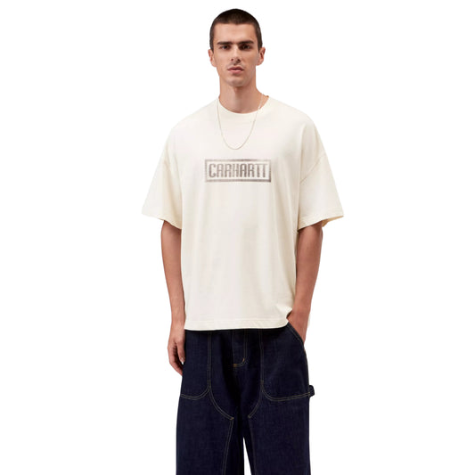 T-shirt Carhartt WIP S/S Bricky Man em Wax Stone Dyed — modelo masculino de frente, meio corpo, cabelo curto, fio de prata fino ao pescoço, gráfico Bricky Man impresso ao centro do peito: logotipo "Carhartt" em caixa retangular com textura de tijolos em cinzento sobre bege creme, malha de algodão orgânico Wax stone-dyed, calças wide-leg denim azul-escuro Carhartt WIP com costuras amarelas, fundo branco.