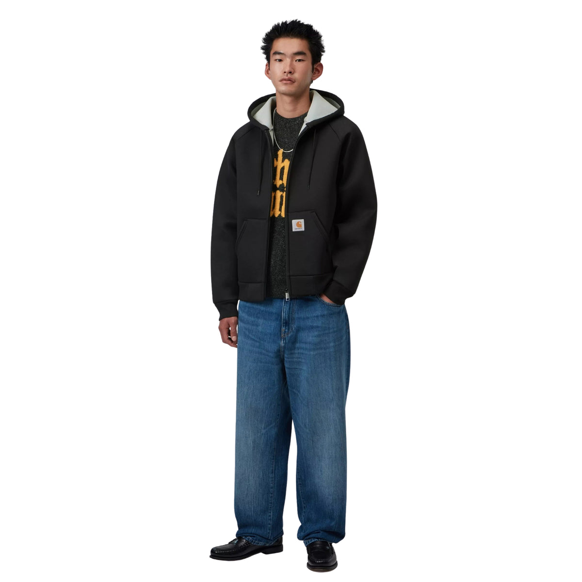 Casaco com carapuço Carhartt WIP Car-Lux em Black/Grey, vista frontal de corpo inteiro no modelo, usado aberto sobre uma t-shirt escura com estampado amarelo, com jeans largos azuis e sapatos pretos.
