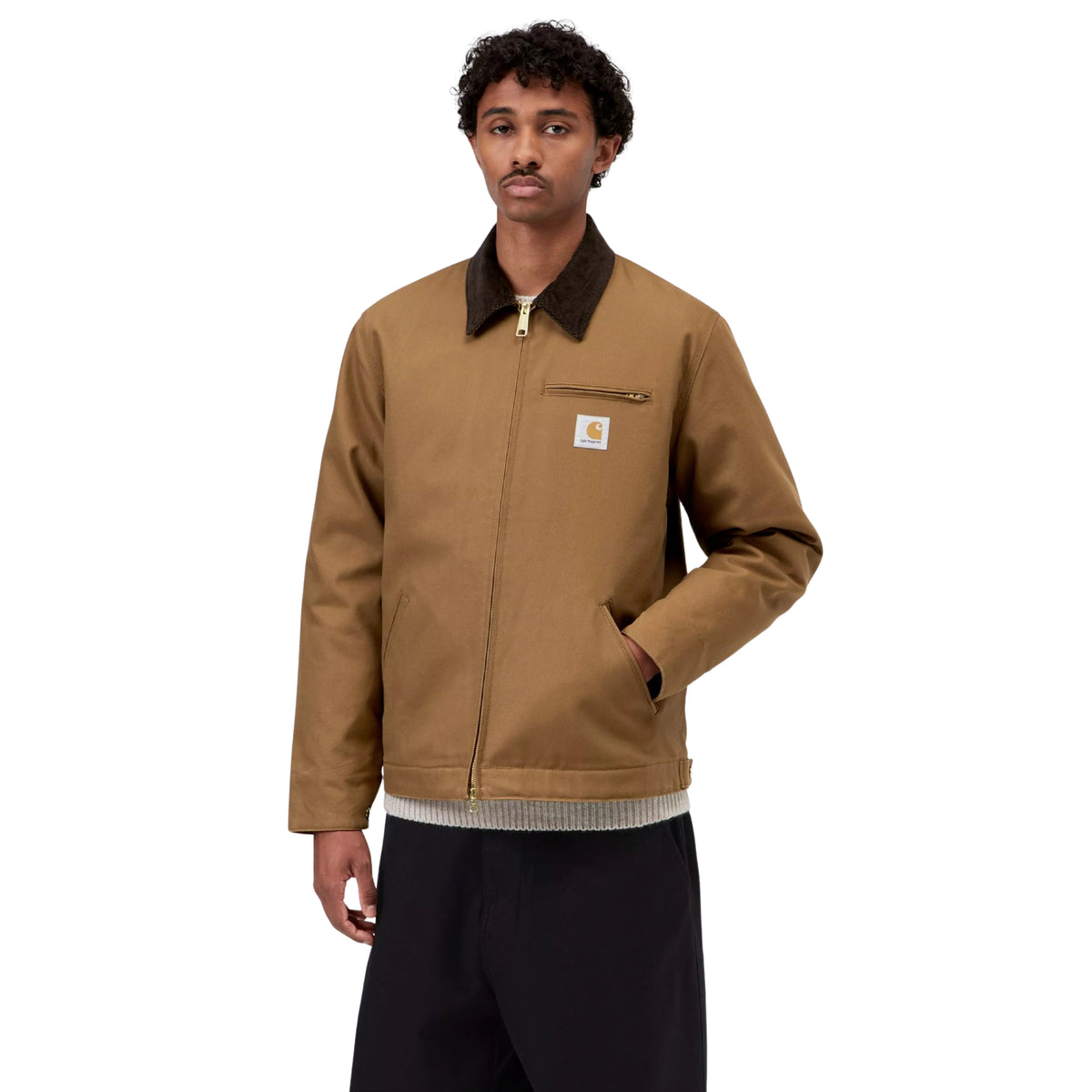 Casaco Carhartt WIP Detroit em Hamilton Brown/Tobacco Rigid, vista frontal em plano médio no modelo, com fecho fechado, gola escura, bolso de peito com fecho e uma mão no bolso lateral.