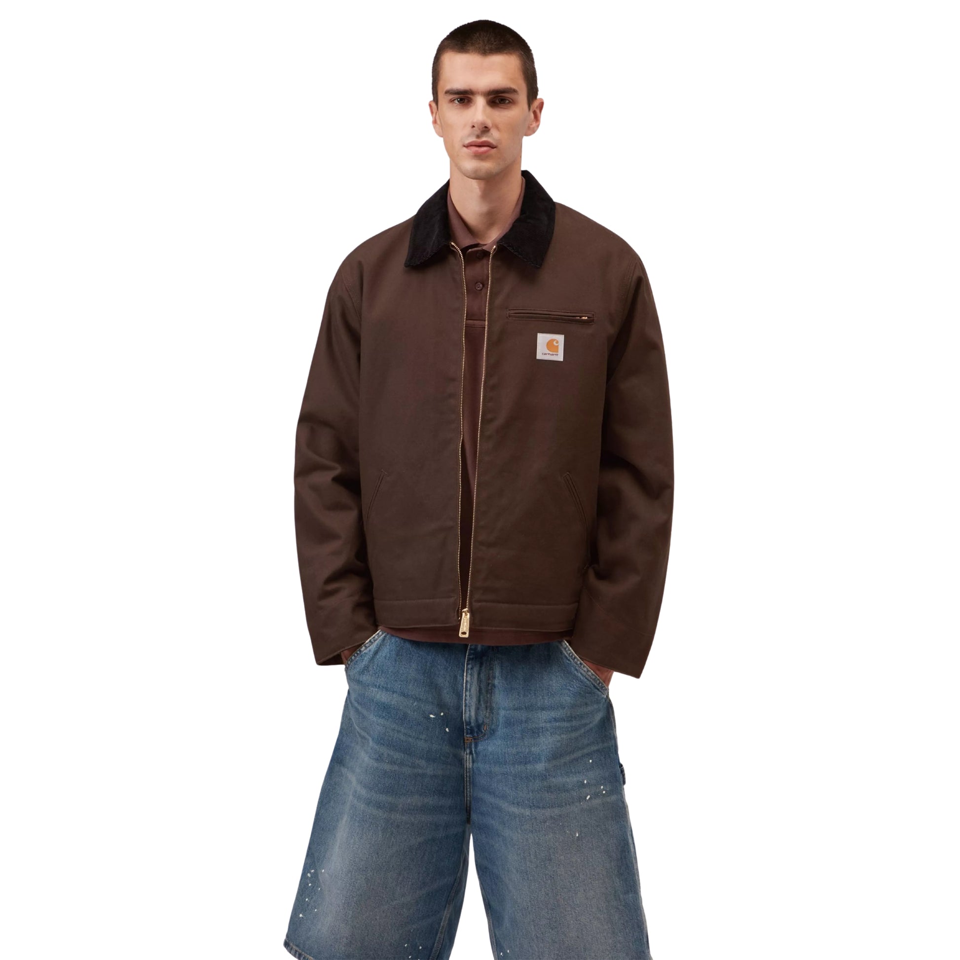 Casaco Carhartt WIP Detroit Jacket em Tobacco / Black Rigid — modelo masculino de frente, meio corpo, calças cargo denim largas azul-claro com respingos de tinta branca, gola em bombazine preta, patch Carhartt WIP no bolso do peito esquerdo e fecho de correr dourado bidirecional visíveis, fundo branco.