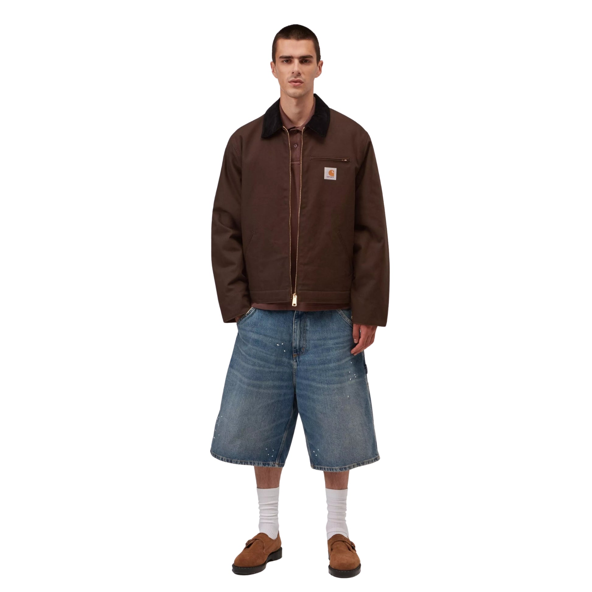Casaco Carhartt WIP Detroit Jacket em Tobacco / Black Rigid — modelo masculino de frente, corpo inteiro, calças cargo denim largas azul-claro com respingos de tinta branca, meias brancas altas e mocassins castanhos, casaco aberto a revelar polo castanho escuro por baixo, fundo branco.