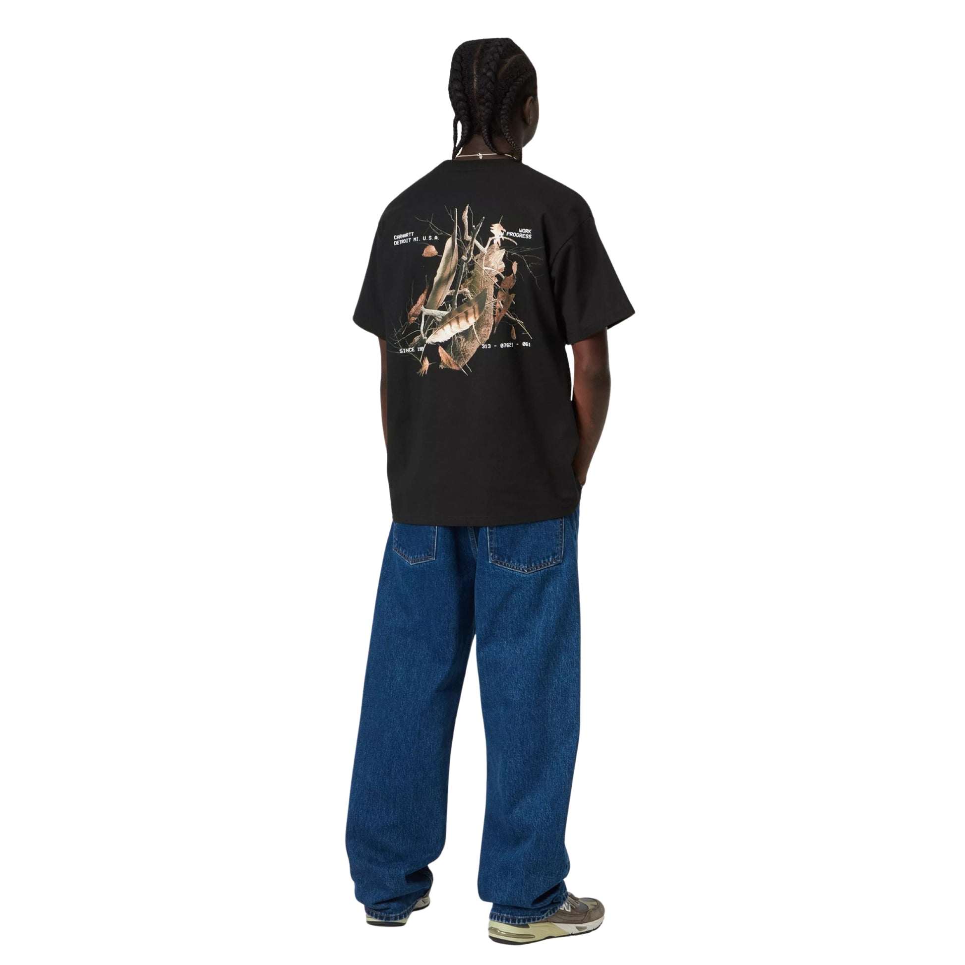 T-shirt Carhartt WIP S/S Feather Progress em Black — modelo masculino de costas, corpo inteiro, cabelo com tranças apanhadas, fio prateado ao pescoço, t-shirt preta de algodão orgânico com print fotográfico de penas e ramos nas costas e texto tipográfico branco Carhartt WIP Detroit, calças wide-leg em ganga azul-escura, sapatilhas cinzentas New Balance, fundo branco.