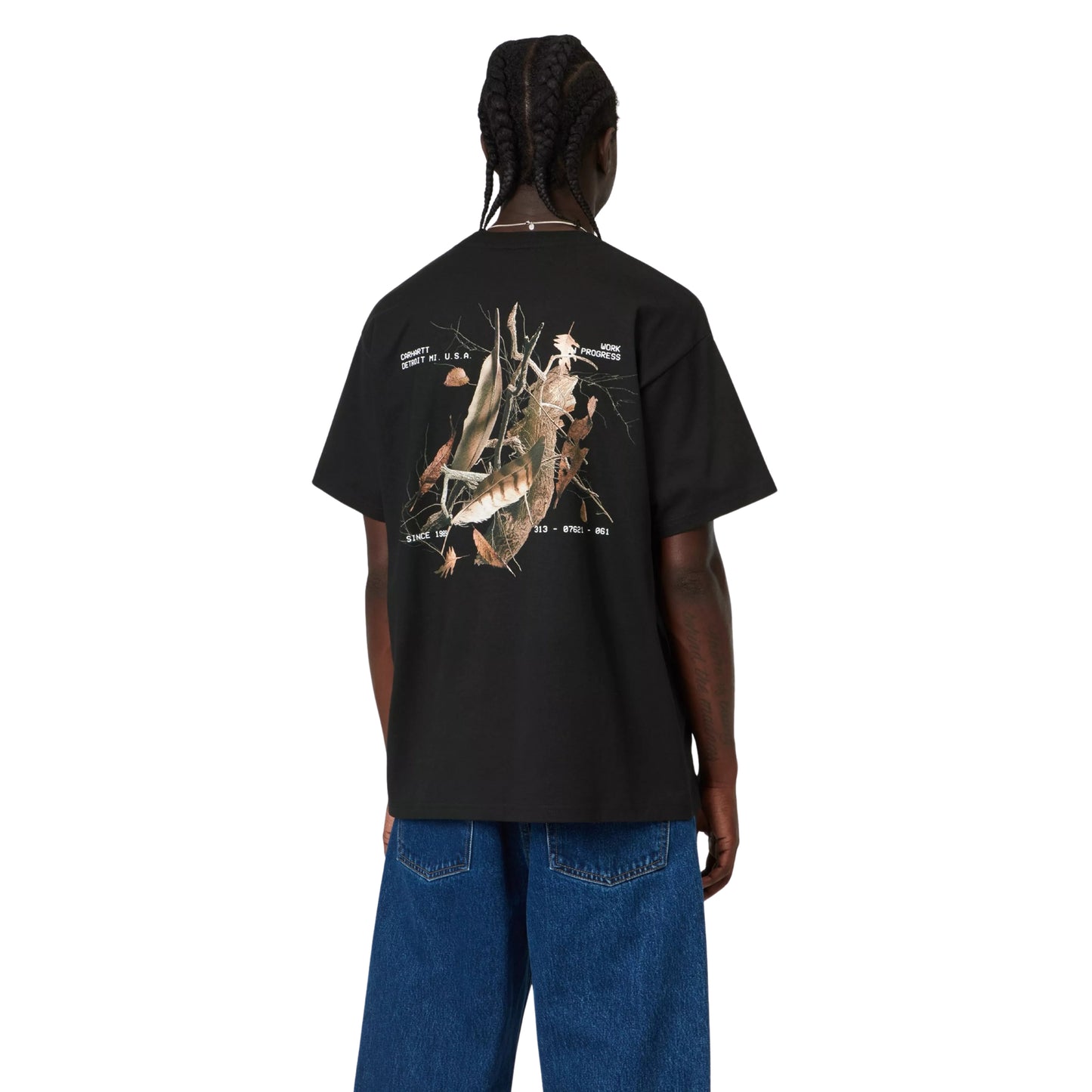 T-shirt Carhartt WIP S/S Feather Progress em Black — modelo masculino de costas, meio corpo superior, cabelo com tranças apanhadas, fio prateado ao pescoço, t-shirt preta com print fotográfico de penas, ramos e folhas secas em tons bege e castanho nas costas e texto branco "CARHARTT DETROIT MI. U.S.A. / WORK PROGRESS / SINCE 1989" em destaque, fundo branco.