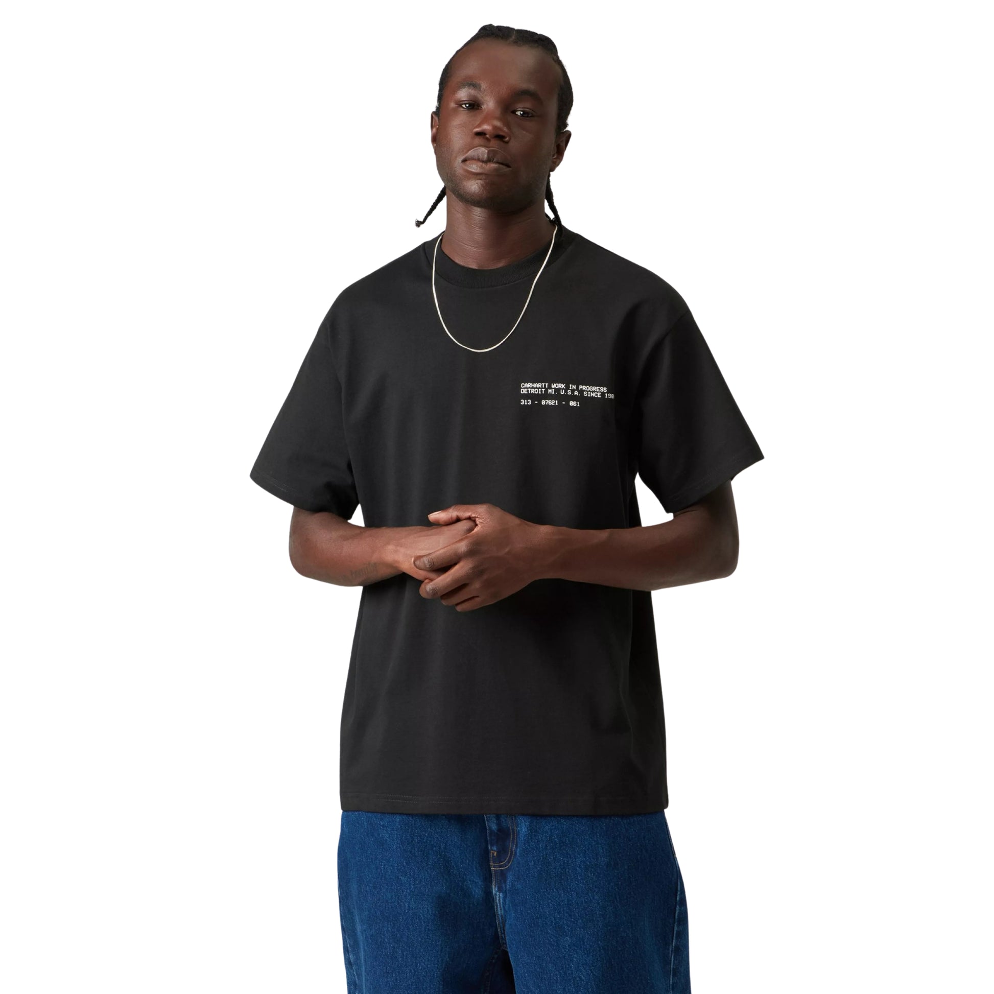 T-shirt Carhartt WIP S/S Feather Progress em Black — modelo masculino de frente, meio corpo superior, cabelo com tranças apanhadas, fio prateado ao pescoço, anel dourado, t-shirt preta de algodão orgânico com corte solto e print de texto branco discreto no peito esquerdo, calças wide-leg em ganga azul, fundo branco.
