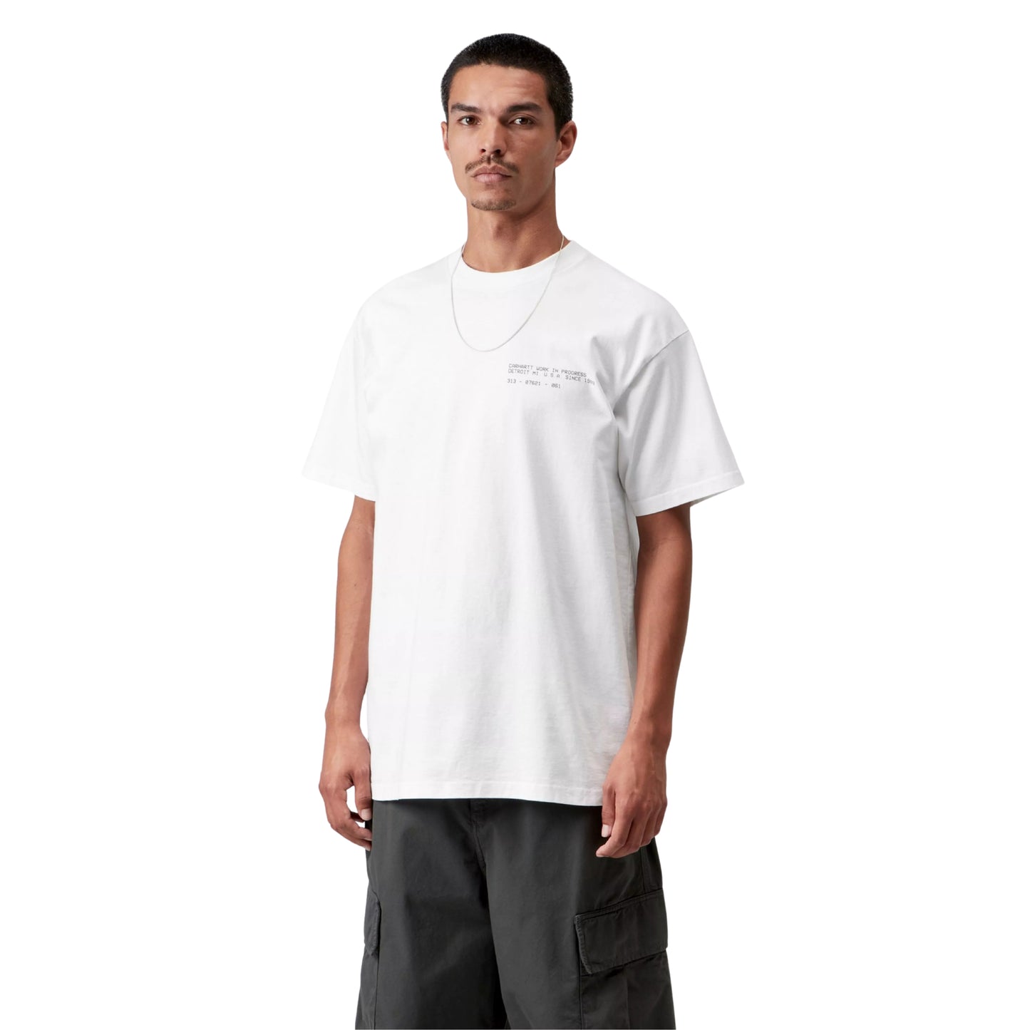 T-shirt Carhartt WIP S/S Feather Progress em White — modelo masculino de frente a 3/4, meio corpo, cabelo curto escuro, bigode, corrente dourada ao pescoço, calças cargo cinzento-escuro Carhartt WIP, texto tipográfico discreto "Carhartt Work In Progress / Detroit MI U.S.A. Since 1989 / 313 – 07621 – 061" estampado no peito esquerdo, decote redondo, corte solto com ombros descaídos, malha de algodão biológico branco, fundo branco.
