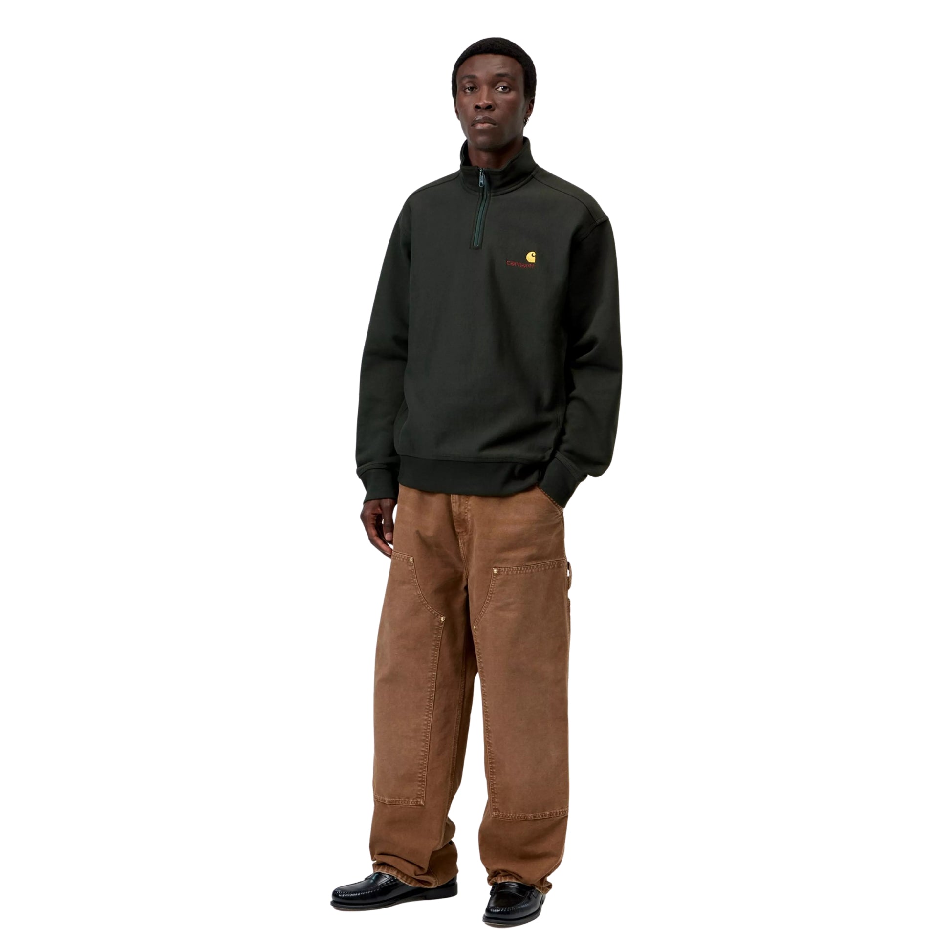 Sweatshirt Carhartt WIP Half Zip American Script em Raven — modelo a usar a peça de frente, corpo inteiro, gola alta com fecho de correr, bordado American Script com logótipo C amarelo no peito esquerdo, calças Double Knee em castanho Hamilton Brown desgastado e mocassins pretos, fundo branco.