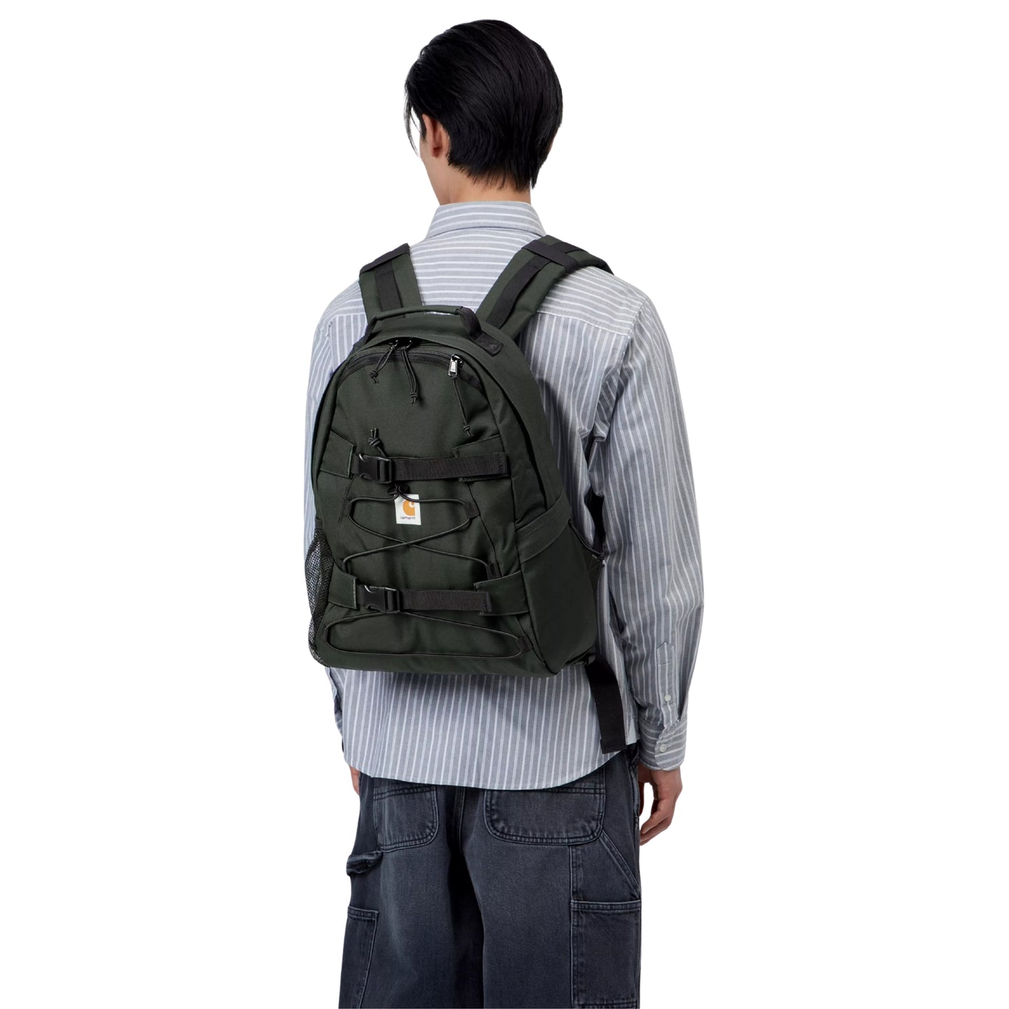 Mochila Carhartt WIP Kickflip Backpack em Raven — modelo de costas com mochila preta às costas, alças acolchoadas ajustáveis, fivelas e fitas elásticas frontais visíveis, combinado com camisa às riscas e calças cargo de ganga.

