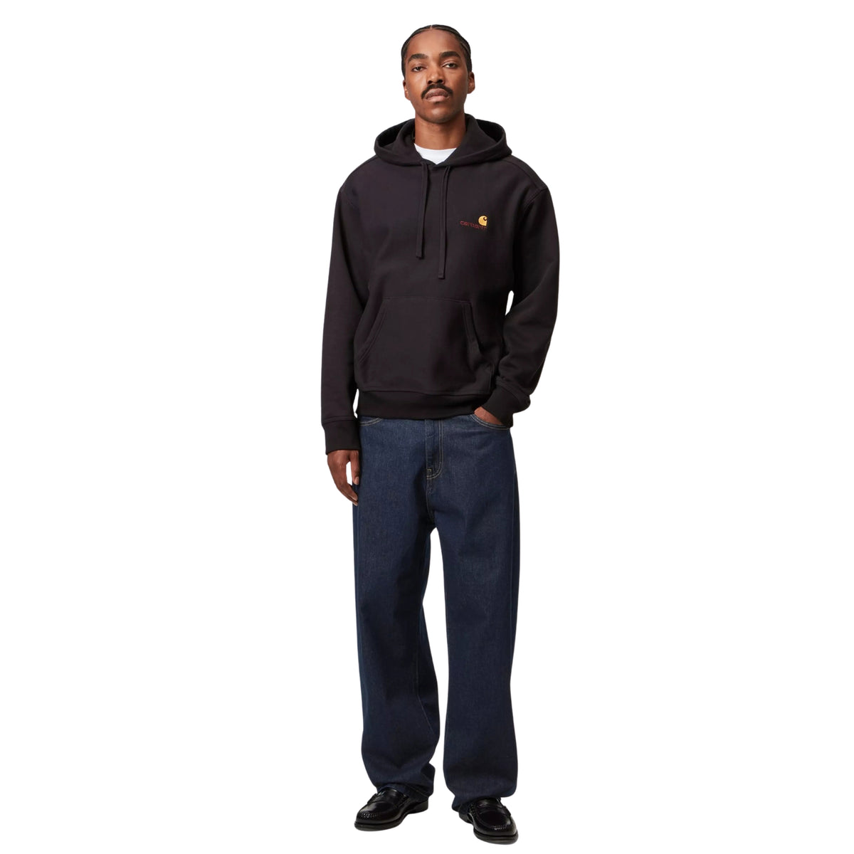 Modelo de corpo inteiro com calças de ganga Carhartt WIP Landon em azul Rinsed, sweat preta com carapuço e sapatos pretos.