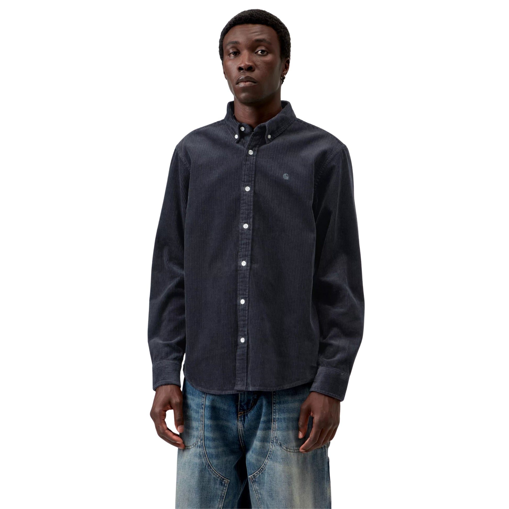 Camisa Carhartt WIP L/S Madison Cord Shirt em Dark Navy — modelo masculino de frente, meio corpo superior, cabelo curto, brinco pequeno, camisa de bombazina azul-marinho escuro com corte regular, colarinho button-down, botões brancos, logótipo "C" bordado em tom-sobre-tom no peito esquerdo, calças wide-leg denim azul-claro com desgaste Carhartt WIP, fundo branco.