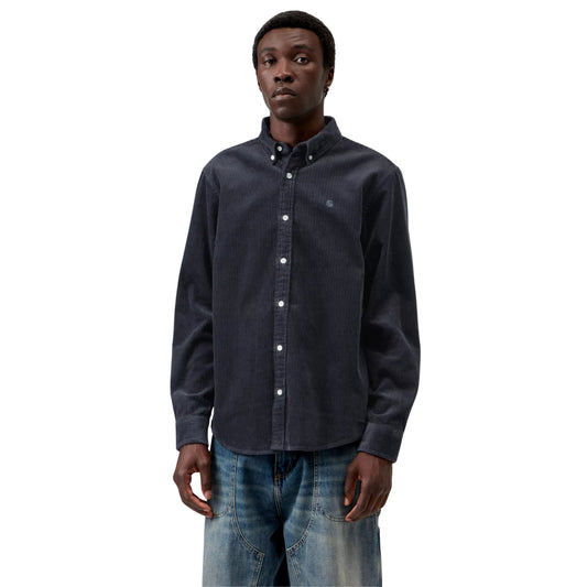 Camisa Carhartt WIP L/S Madison Cord Shirt em Dark Navy — modelo masculino de frente, meio corpo superior, cabelo curto, brinco pequeno, camisa de bombazina azul-marinho escuro com corte regular, colarinho button-down, botões brancos, logótipo "C" bordado em tom-sobre-tom no peito esquerdo, calças wide-leg denim azul-claro com desgaste Carhartt WIP, fundo branco.