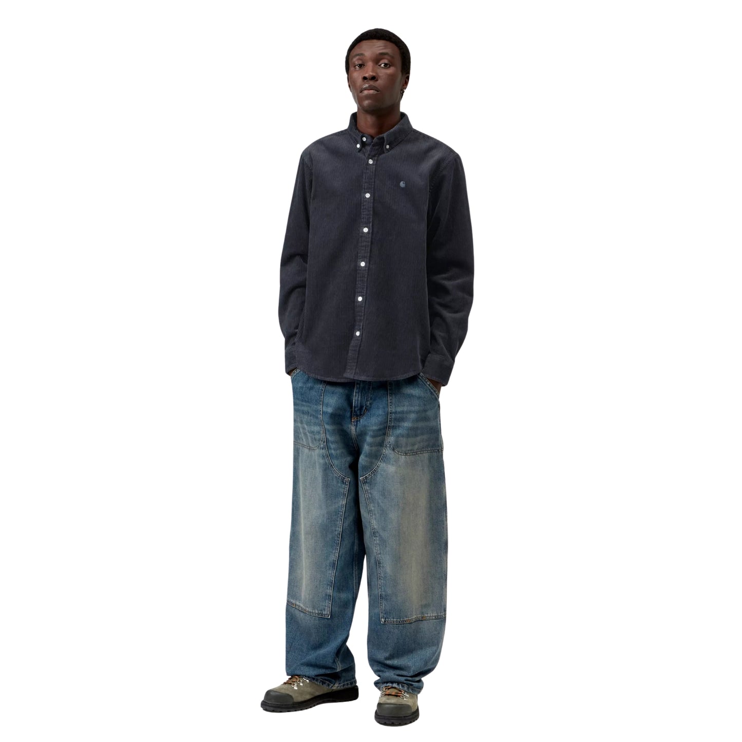 Camisa Carhartt WIP L/S Madison Cord Shirt em Dark Navy — modelo masculino de corpo inteiro, cabelo curto, brinco pequeno, camisa de bombazina azul-marinho escuro com corte regular e barra curva, mãos nos bolsos, calças wide-leg denim azul-claro Double Knee com desgaste Carhartt WIP, botas de caminhada cinzentas, fundo branco.