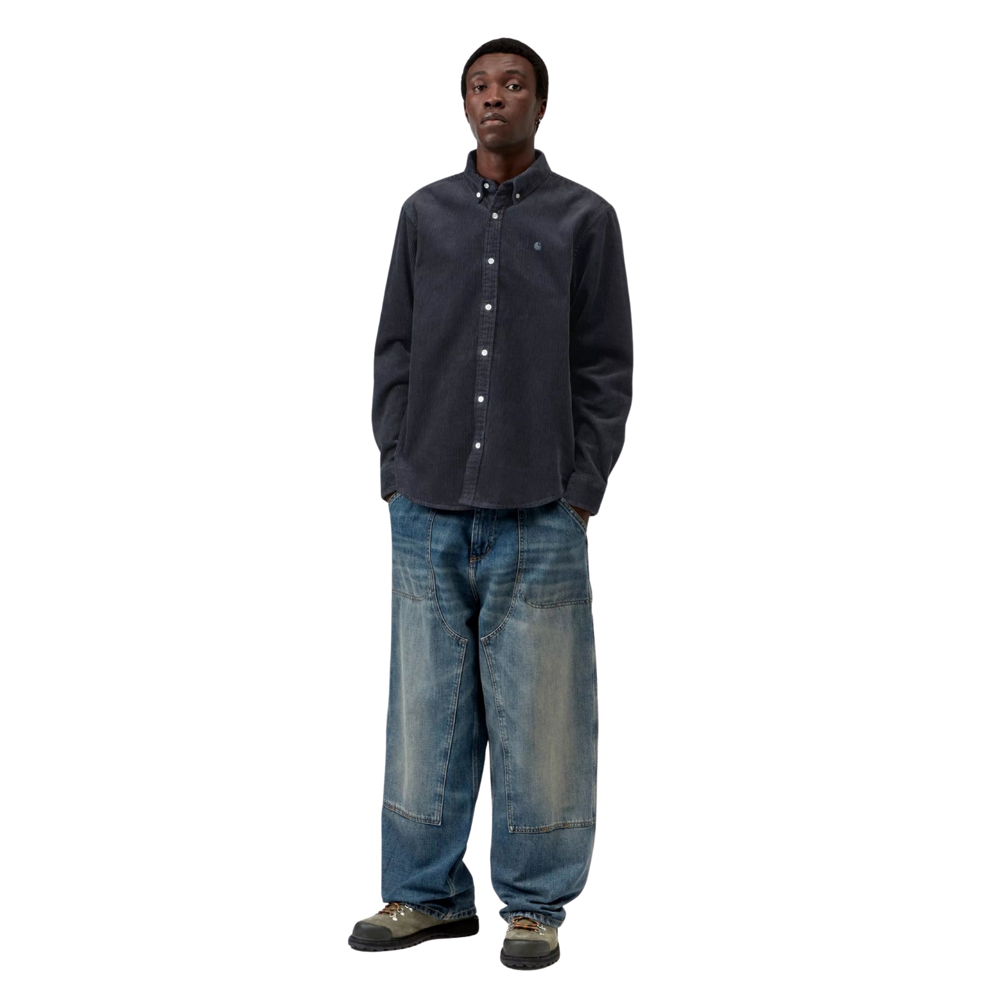 Camisa Carhartt WIP L/S Madison Cord Shirt em Dark Navy — modelo masculino de corpo inteiro, cabelo curto, brinco pequeno, camisa de bombazina azul-marinho escuro com corte regular e barra curva, mãos nos bolsos, calças wide-leg denim azul-claro Double Knee com desgaste Carhartt WIP, botas de caminhada cinzentas, fundo branco.