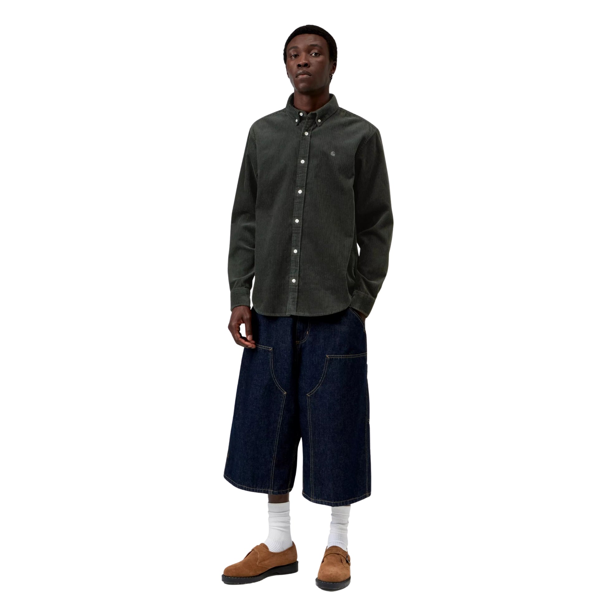 Camisa Carhartt WIP L/S Madison Cord Shirt em Raven — modelo masculino de frente, corpo inteiro, cabelo preto curto afro, camisa de bombazina verde-floresta muito escuro com corte regular, colarinho button-down, botões brancos, logótipo "C" bordado em tom-sobre-tom no peito esquerdo, calças wide-leg carpenter denim azul-escuro com costuras contrastantes brancas Carhartt WIP, meias brancas altas, sapatos derby em camurça castanha, fundo branco.