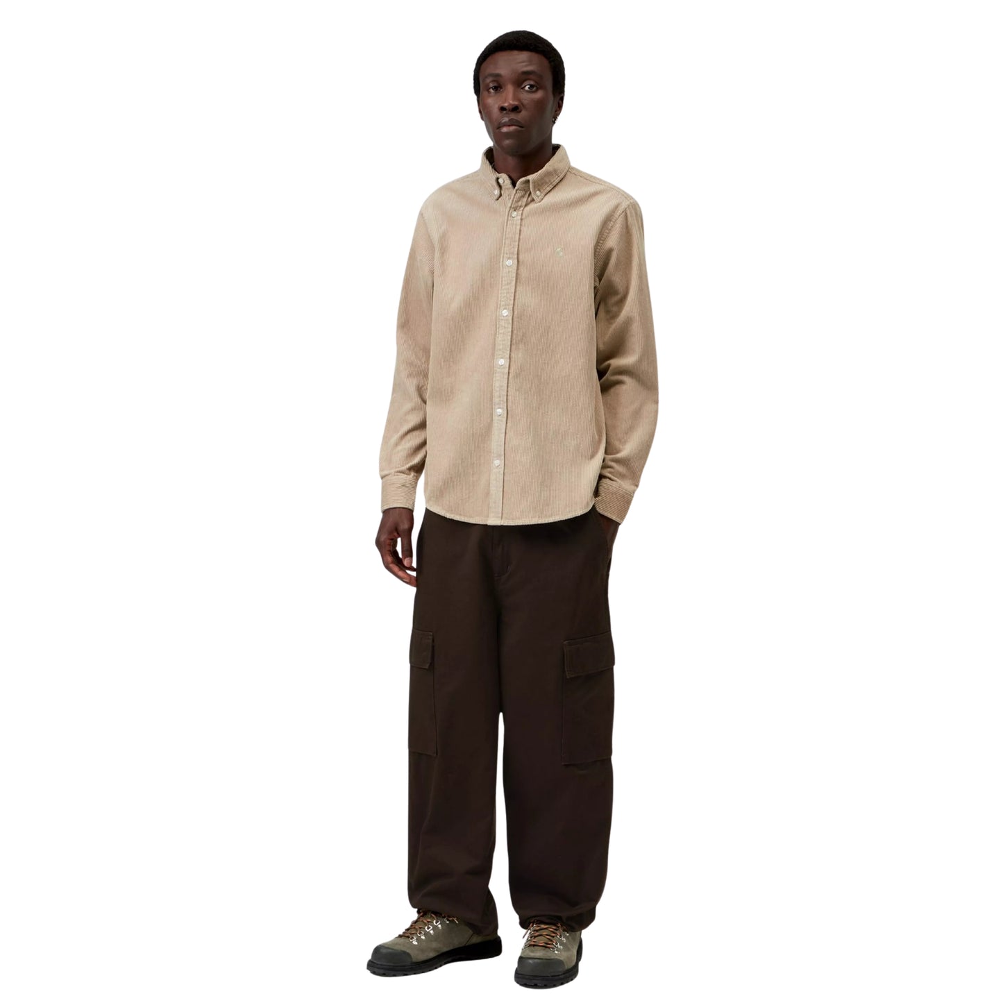 Camisa Carhartt WIP L/S Madison Cord Shirt em Wall — modelo masculino de corpo inteiro, cabelo curto preto, brinco pequeno, camisa de bombazina bege-areia com corte regular e logótipo "C" em tom-sobre-tom no peito esquerdo, colarinho button-down, botões brancos, calças cargo wide-leg castanho-escuro Carhartt WIP, botas de caminhada cinzentas, fundo branco.
