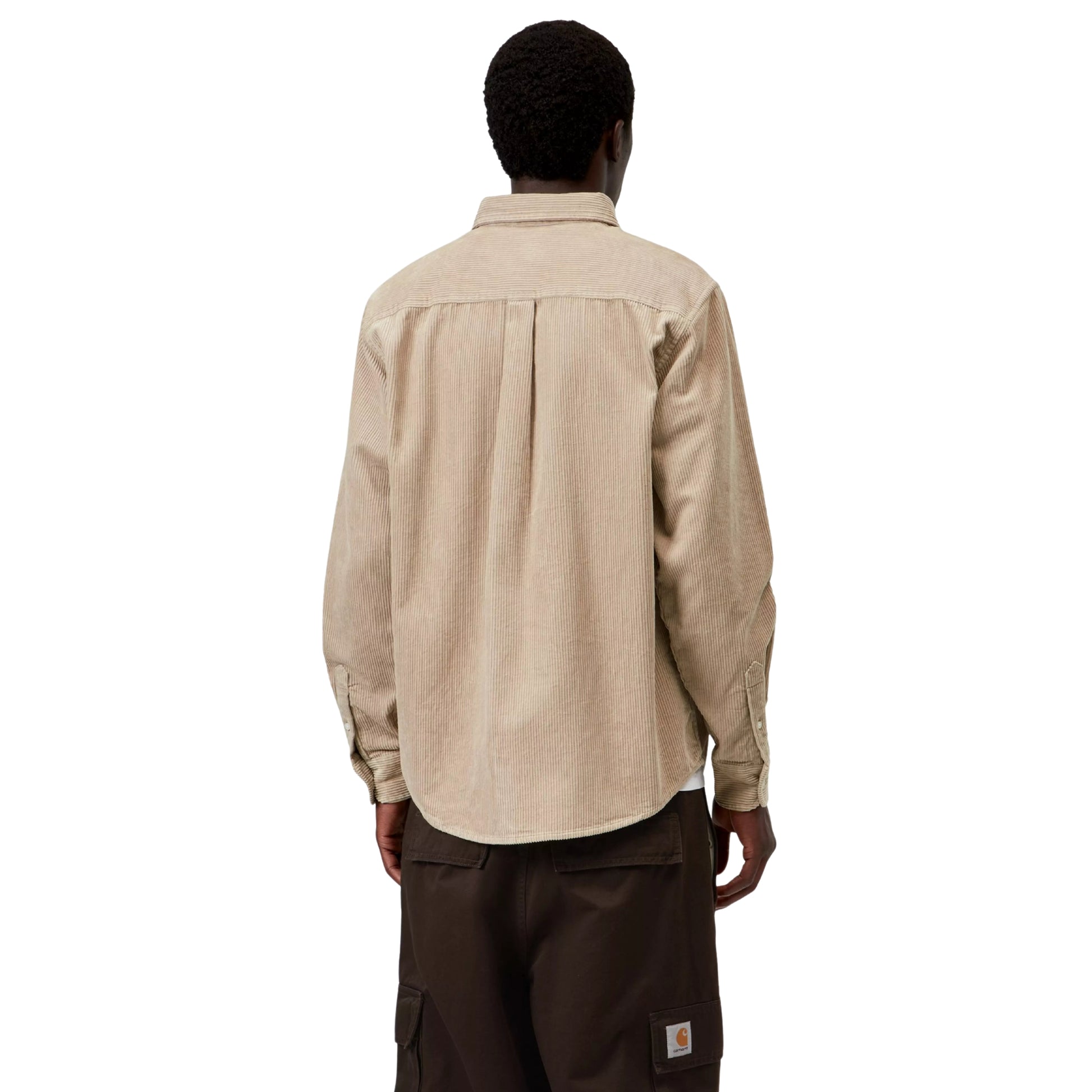 Camisa Carhartt WIP L/S Madison Cord Shirt em Wall — modelo masculino de costas, meio corpo superior e inferior, cabelo curto preto, camisa de bombazina bege-areia com corte regular, pala central nas costas, canelado largo, punho com botão, barra curva, calças cargo wide-leg castanho-escuro com Square Label Carhartt WIP dourado visível no bolso traseiro, fundo branco.