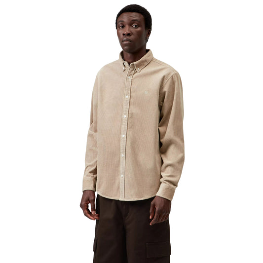 Camisa Carhartt WIP L/S Madison Cord Shirt em Wall — modelo masculino de frente, meio corpo superior, cabelo curto preto, brinco pequeno, camisa de bombazina bege-areia com colarinho button-down, fecho com botões brancos, logótipo "C" Carhartt bordado em tom-sobre-tom no peito esquerdo em destaque, canelado largo da bombazina visível, calças cargo castanho-escuro, fundo branco.