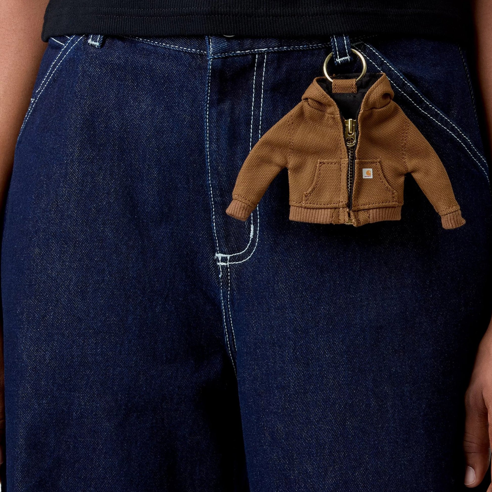Porta-chaves Carhartt WIP Mini Active Jacket Keychain em Hamilton Brown — detalhe de uso, berloque preso ao passante de jeans wide-leg azul-escuro Carhartt WIP com anel metálico dourado, vista frontal da miniatura da Active Jacket em lona Dearborn castanha Hamilton Brown com fecho dourado e etiqueta woven dourada no bolso, fundo de tecido denim com costuras brancas.