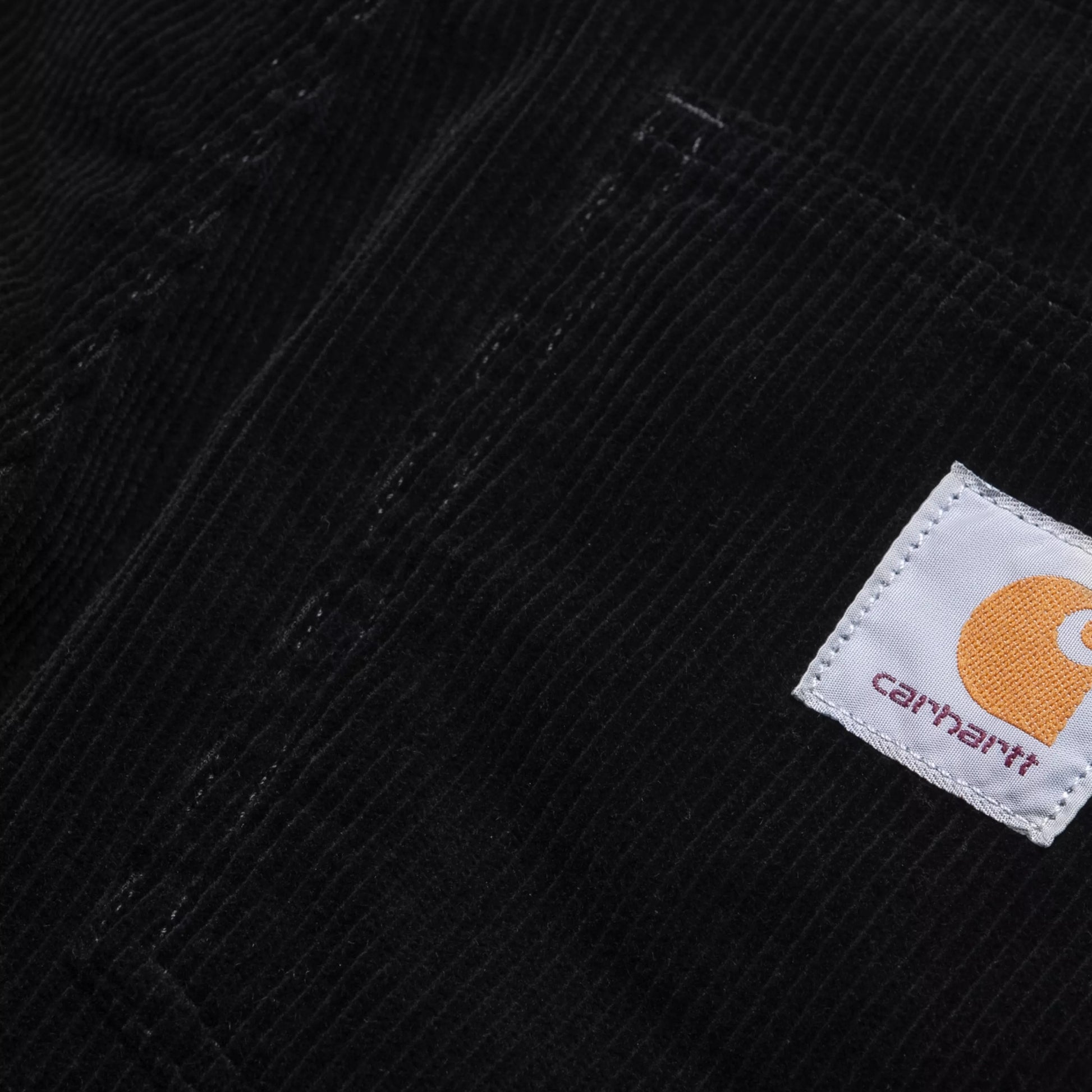 Calças Carhartt WIP Newel Pant em Black — detalhe da cintura traseira em close, bombazina Coventry de oito nervuras preta, Square Label Carhartt WIP em tecido branco com "C" laranja e texto "carhartt" em roxo costurada no bolso traseiro esquerdo, textura canelada larga em destaque, fundo branco.