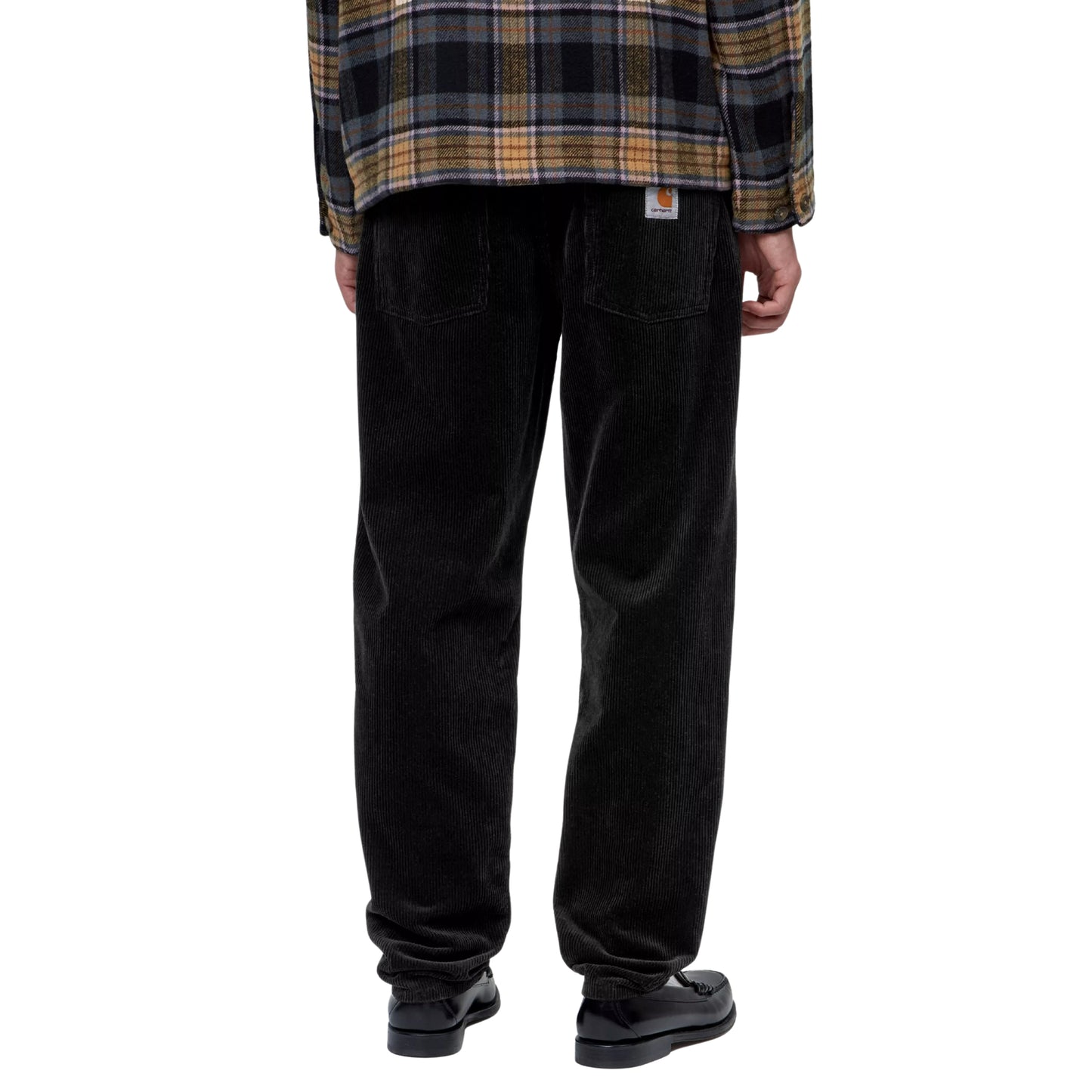 Calças Carhartt WIP Newel Pant em Black — modelo masculino de costas, meio corpo inferior, casaco xadrez oversized Carhartt WIP, calças em bombazina Coventry preta com corte relaxado afunilado, dois bolsos traseiros, Square Label Carhartt WIP branca com "C" laranja na cintura traseira, loafers pretos, fundo branco.