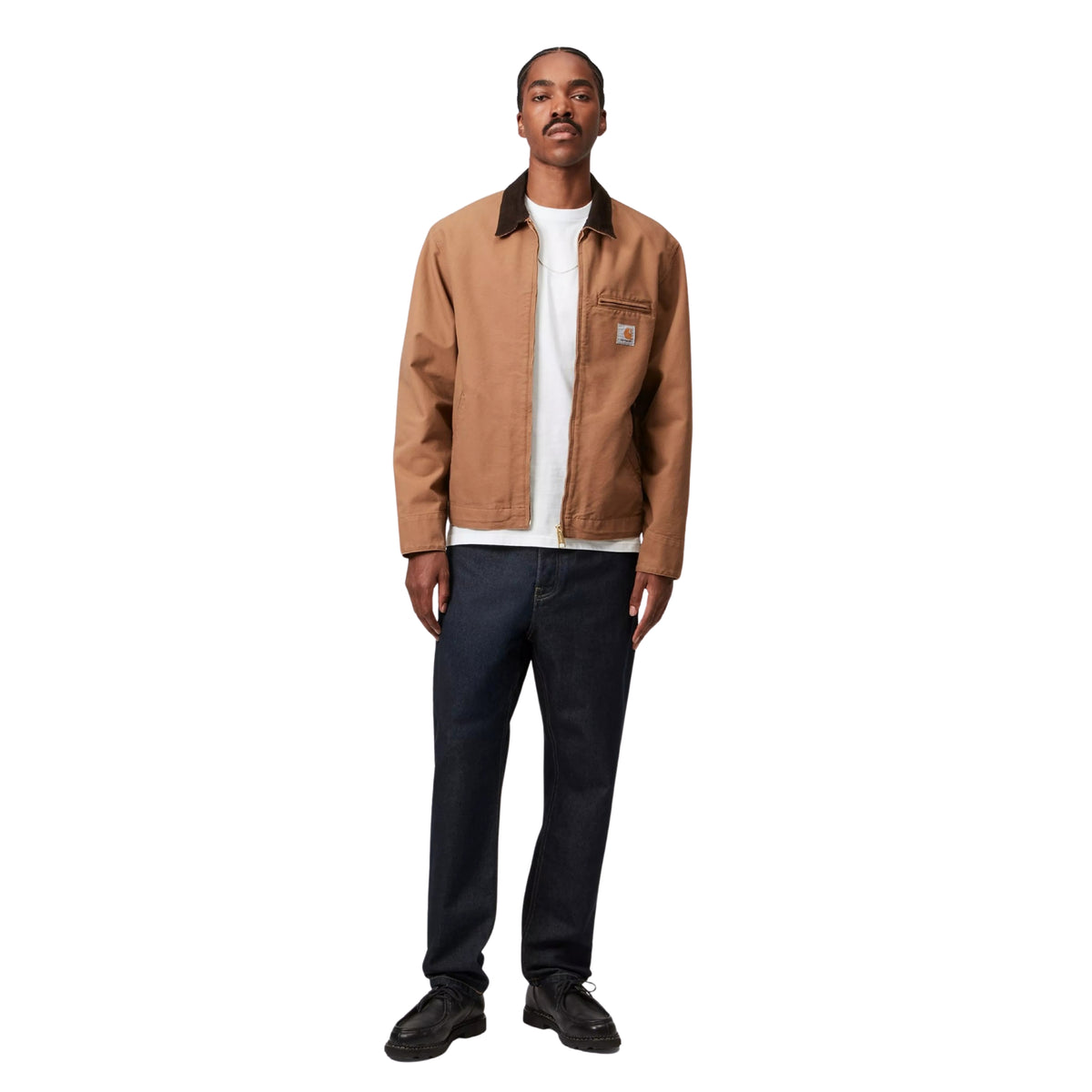 Modelo de corpo inteiro com calças de ganga Carhartt WIP Newel em azul One Wash, combinadas com casaco castanho, t-shirt branca e sapatos pretos.