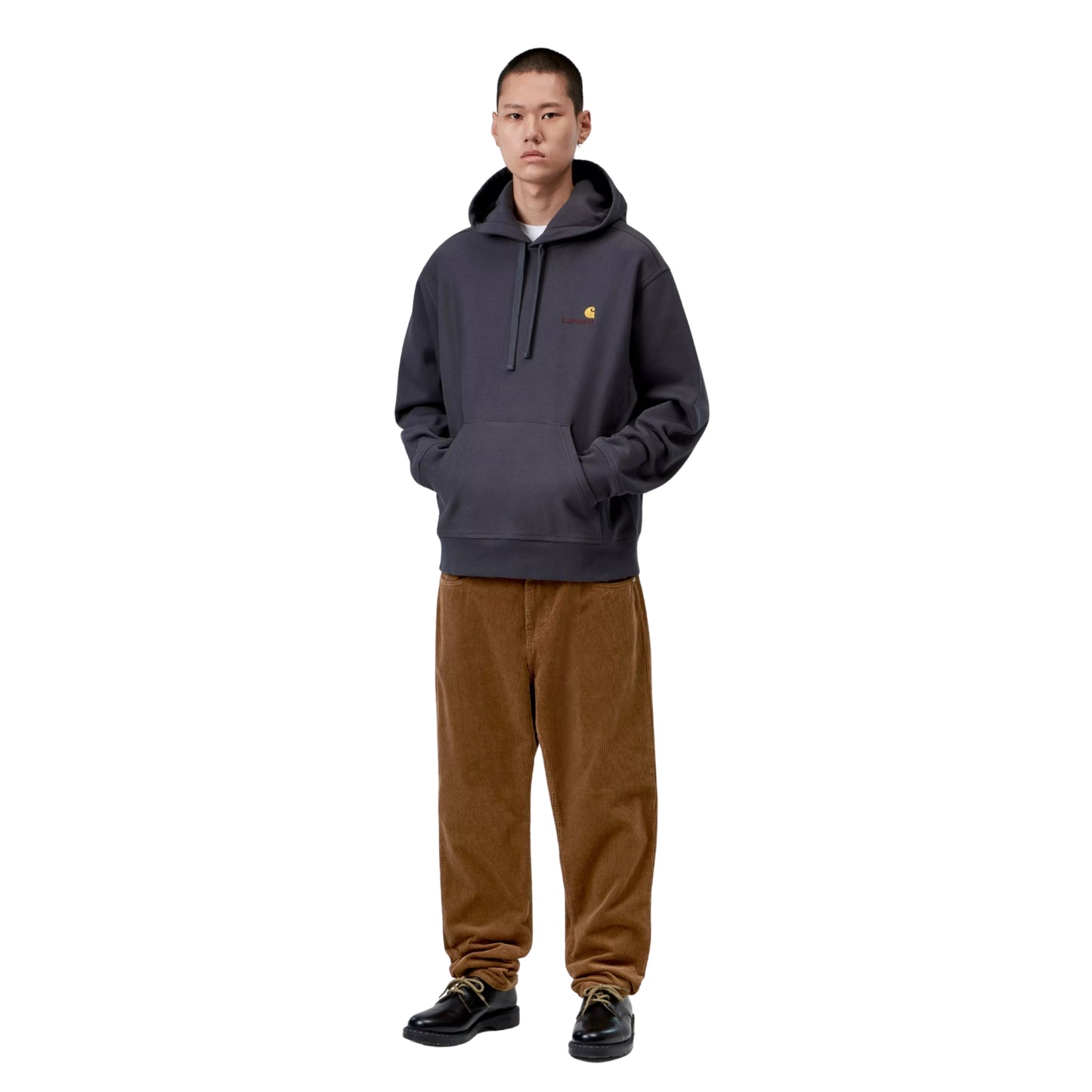 Calças Carhartt WIP Newel Pant em Hamilton Brown Rinsed — modelo masculino asiático de corpo inteiro, cabelo curto preto, sweatshirt com capuz cinzento-azulado Carhartt WIP, calças em bombazina castanha Hamilton Brown rinsed com corte relaxado afunilado a empilhar ligeiramente nas sapatilhas, sapatos pretos de sola plana com atacadores bege, fundo branco.