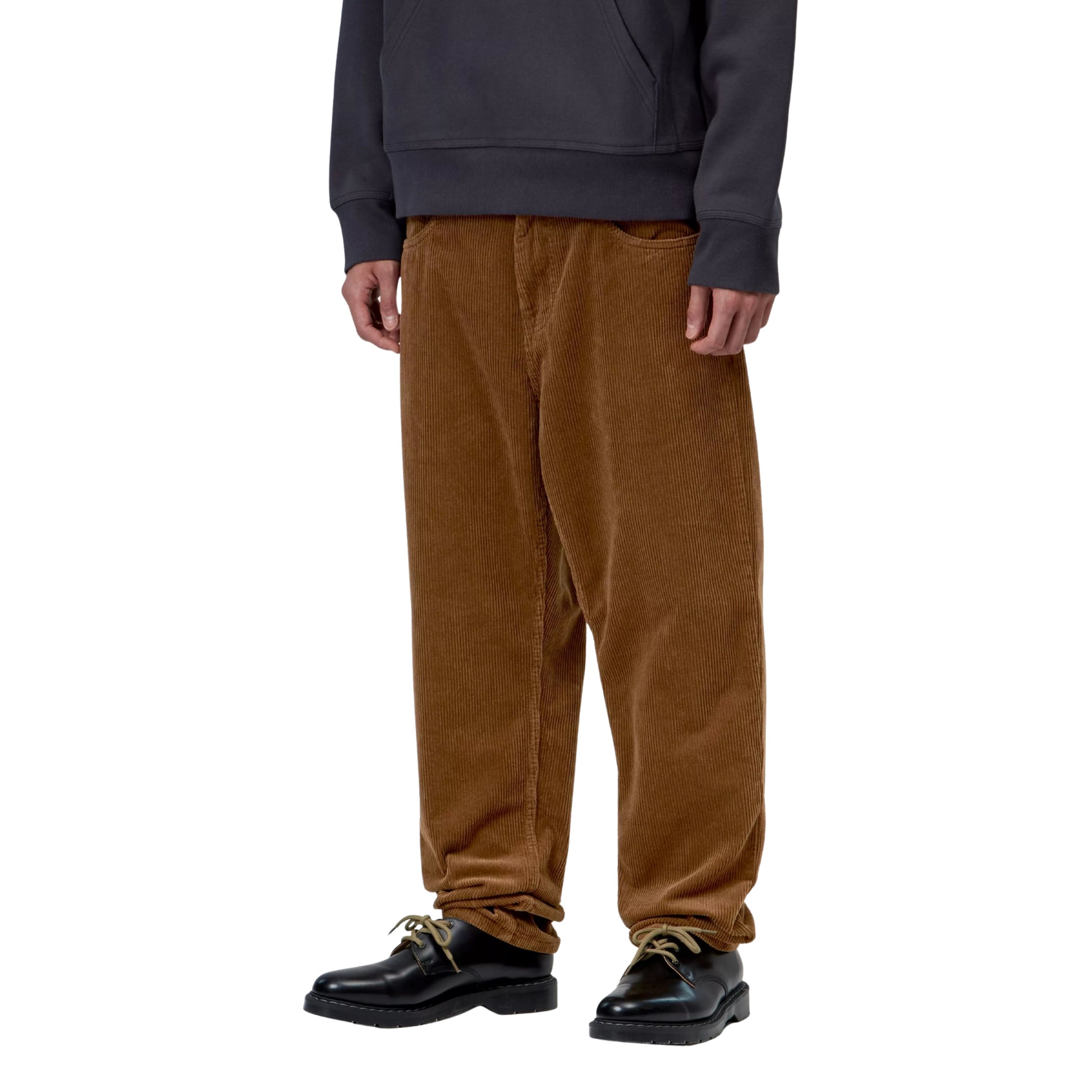Calças Carhartt WIP Newel Pant em Hamilton Brown Rinsed — modelo masculino de frente, meio corpo inferior, sweatshirt com capuz cinzento-azulado, calças em bombazina Coventry castanha Hamilton Brown rinsed, corte relaxado afunilado com cintura regular e bolsos frontais visíveis, sapatos pretos de sola plana, fundo branco.