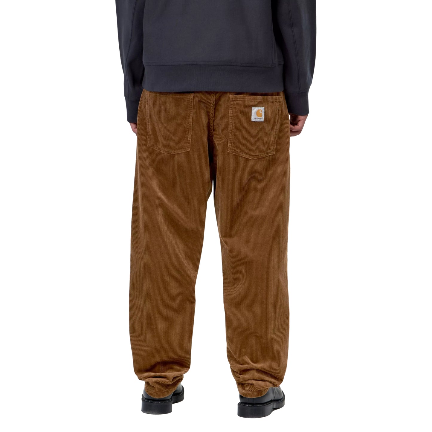 Calças Carhartt WIP Newel Pant em Hamilton Brown Rinsed — modelo masculino de costas, meio corpo inferior, sweatshirt com capuz cinzento-azulado, costas das calças em bombazina Coventry castanha Hamilton Brown rinsed, dois bolsos traseiros, Square Label Carhartt WIP em tecido branco com logotipo dourado no bolso traseiro direito, sapatos pretos de sola plana, fundo branco.